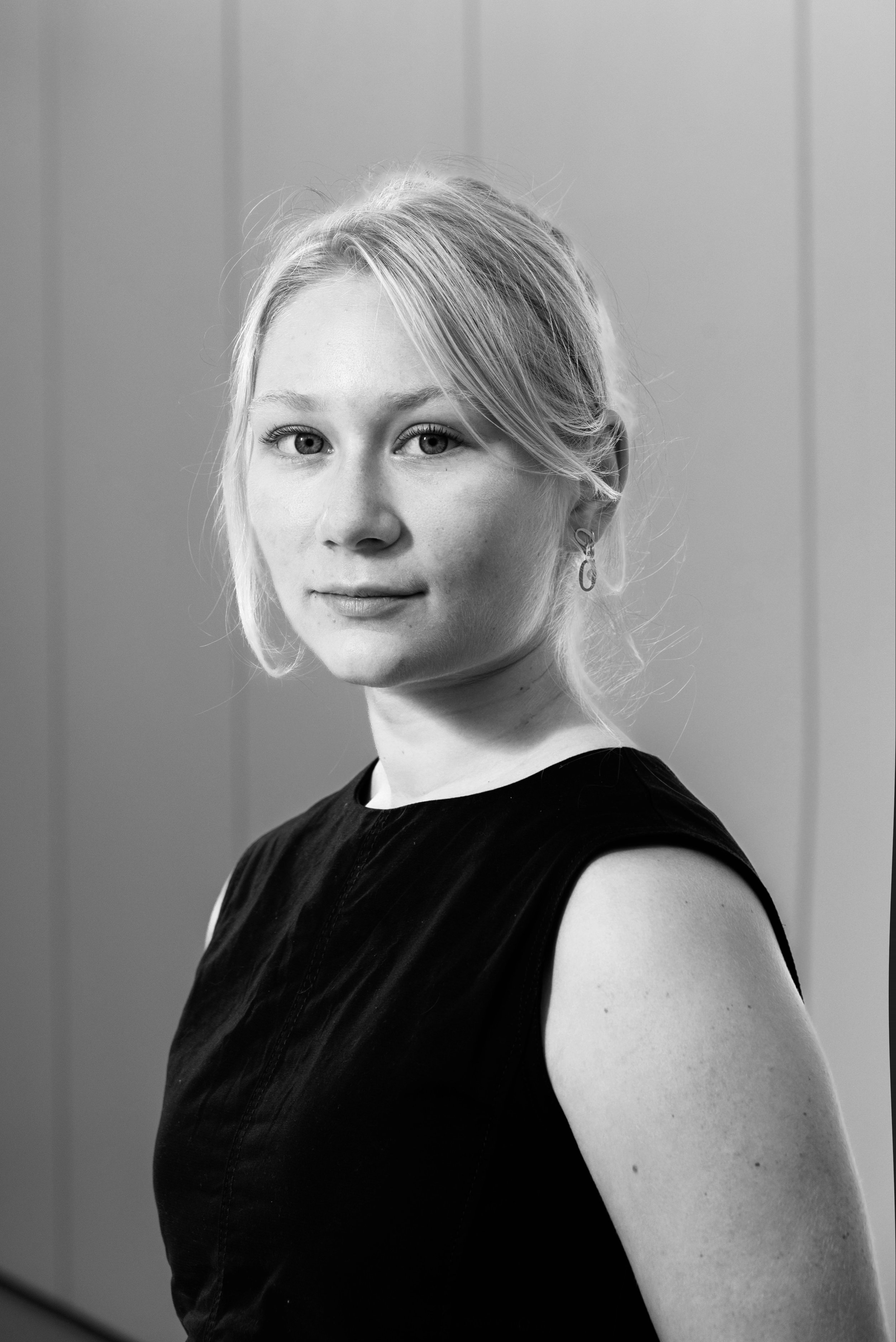 Black and white photo of a young woman with light-colored hair, wearing a sleeveless top, standing against a plain background.