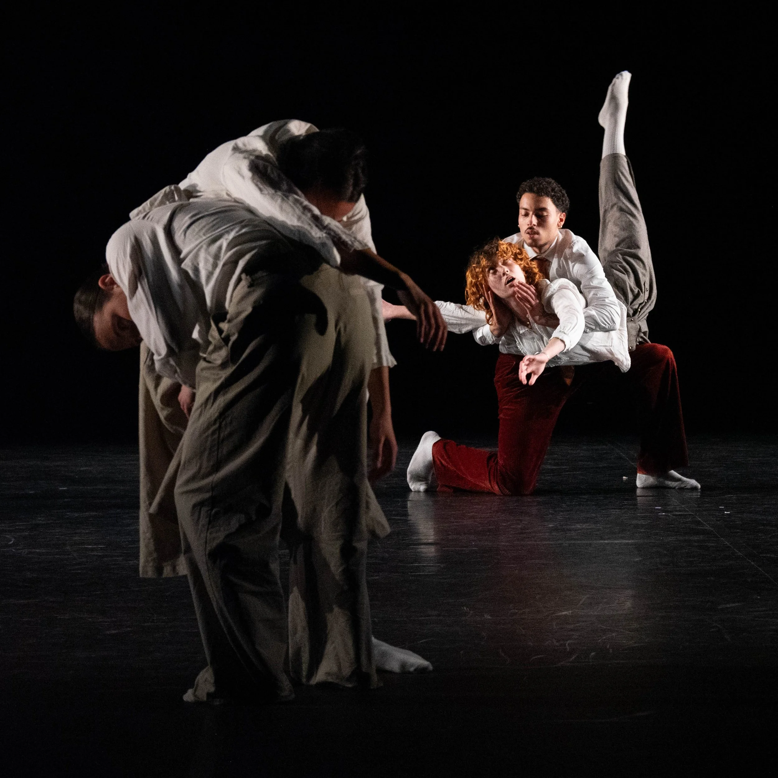 In this stage performance, four performers dressed in white clothing are engaged in a contemporary dance or theatrical act on a dark stage. One performer is in a handstand with one leg extended upwards, another appears to be leaning forward while supporting a child with curly red hair on her lap, and the others are bent or crouched in dynamic poses, all displaying coordinated movements.
