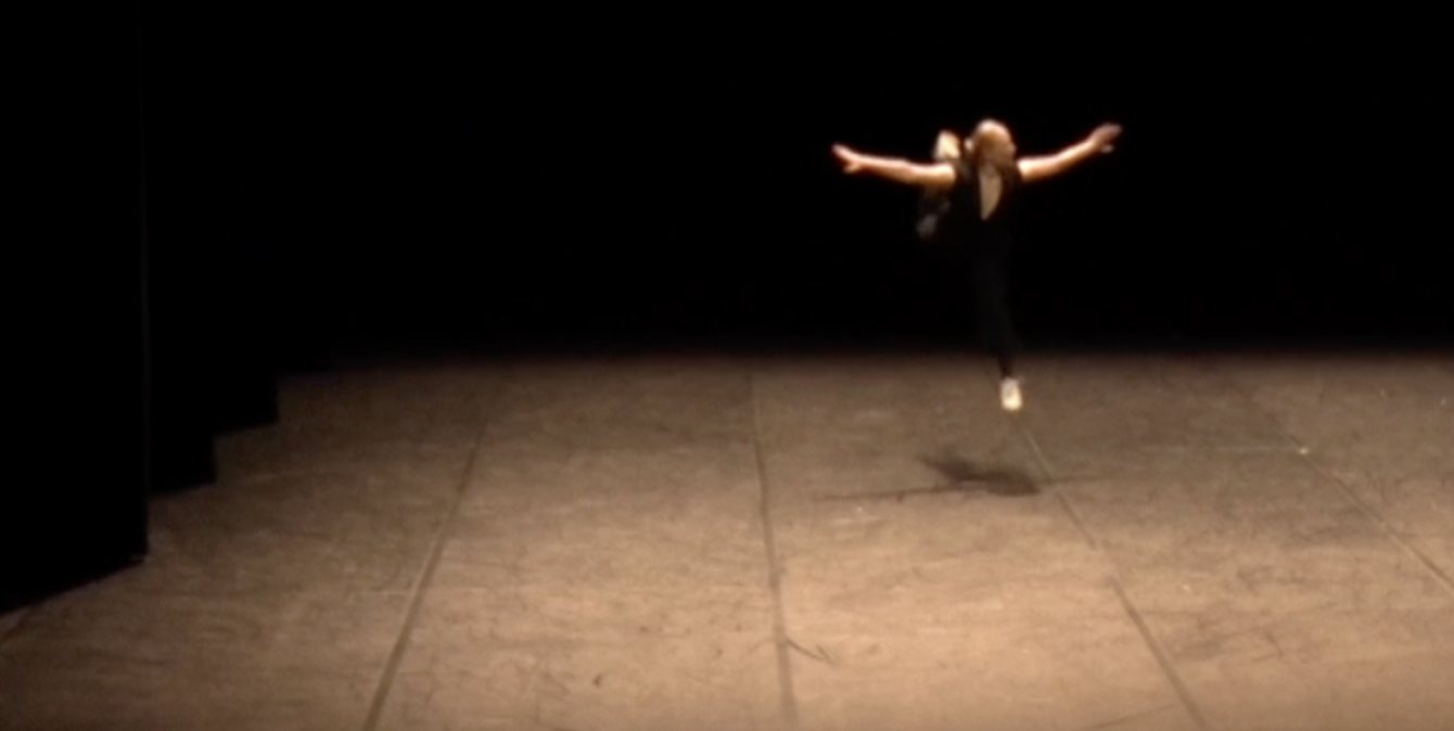 A woman dancing alone on a stage with a dark background, illuminated by a spotlight.