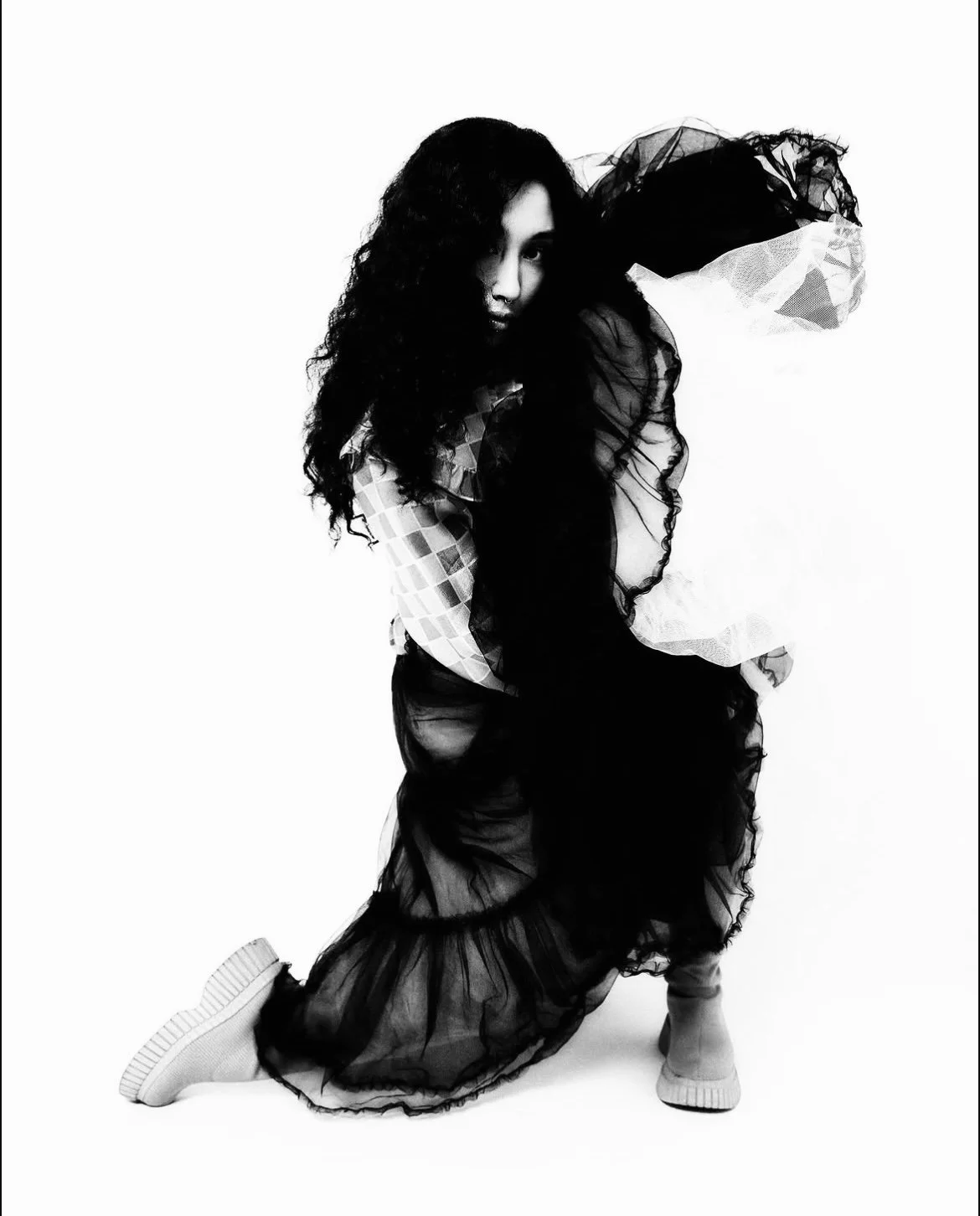 A woman with long curly hair wearing layered black tulle skirt, mesh top, and sneakers in a studio with a plain white background.