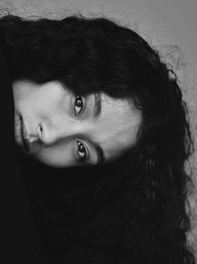 A close-up black and white portrait of a woman with dark curly hair, lying down and looking directly at the camera.