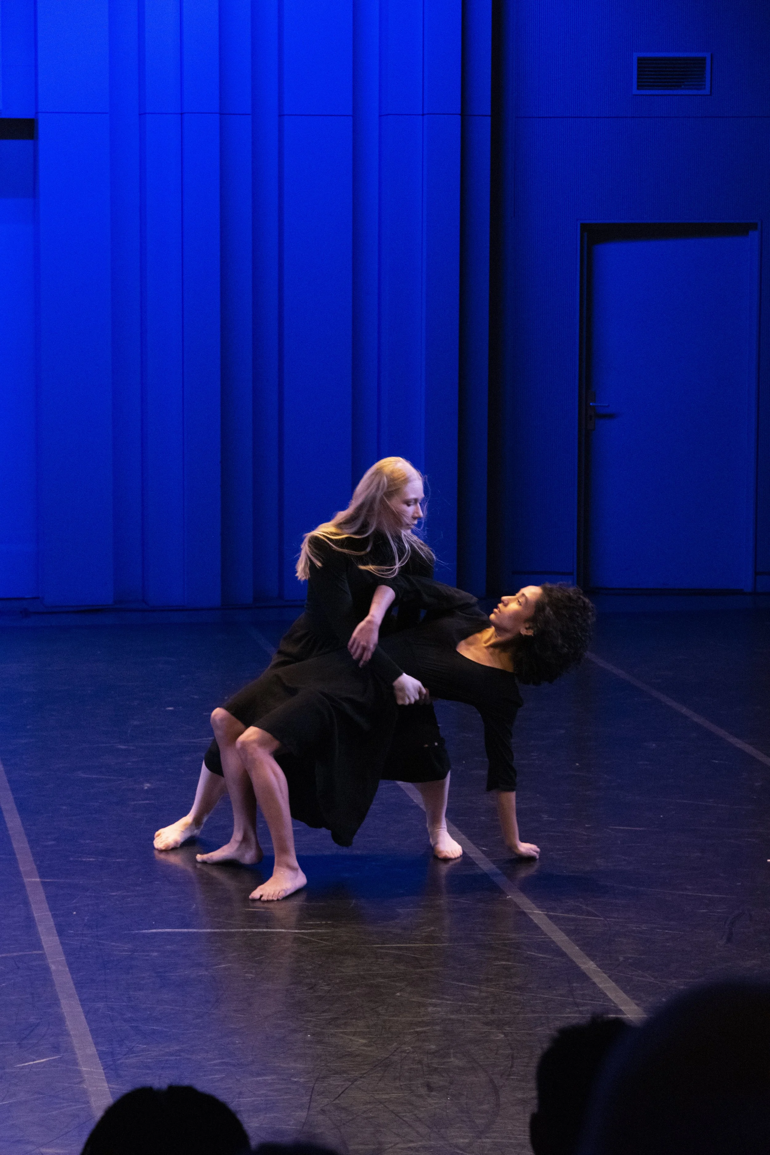 Two women performing contemporary dance on a dark stage with a blue background, one lying on the floor and the other sitting on her back, engaged in a dance routine.