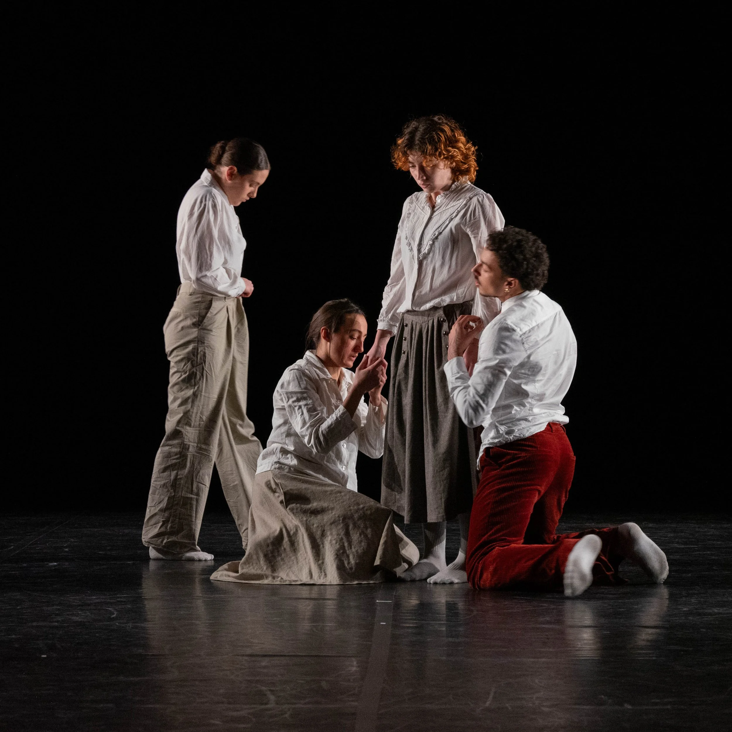 People, wearing white shirts and red pants, gathered closely in a dark stage setting, engaging in a moment of connection.