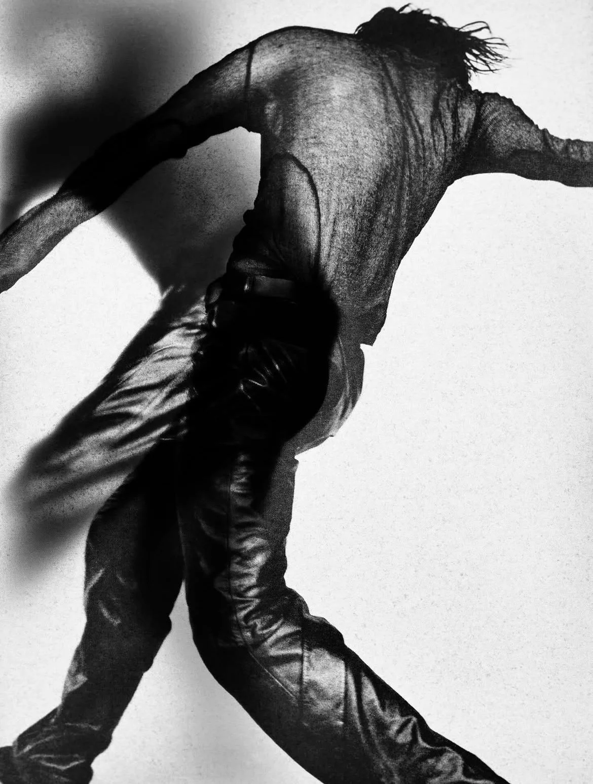 Black and white photo of a person with long hair in a dynamic pose, wearing a semi-sheer top and shiny pants, leaning backward with arms and legs extended.