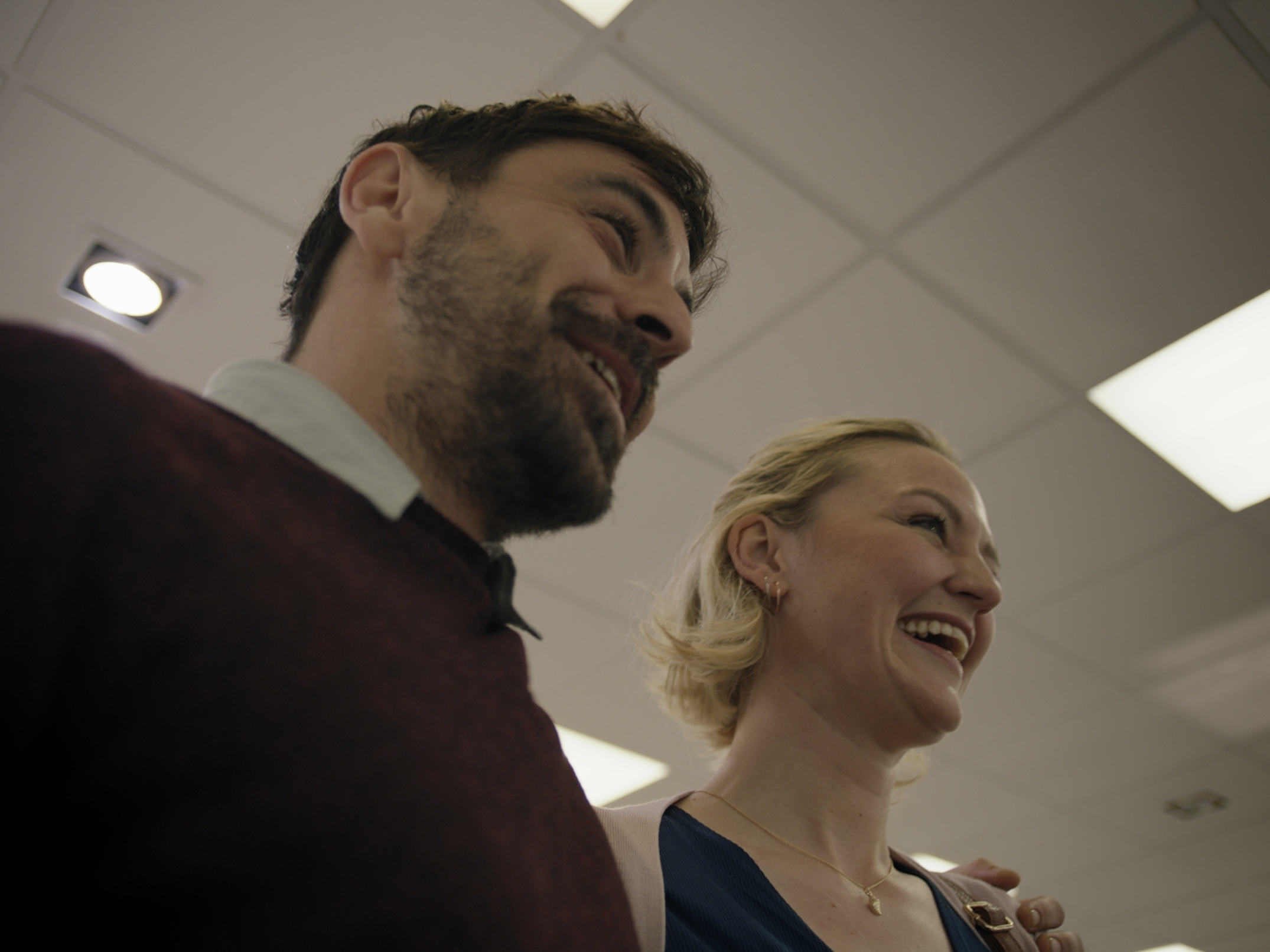 A man and a woman standing side by side with their eyes closed, smiling and appearing to engage in a moment of mindfulness or meditation in an indoor setting with a ceiling and lighting visible.