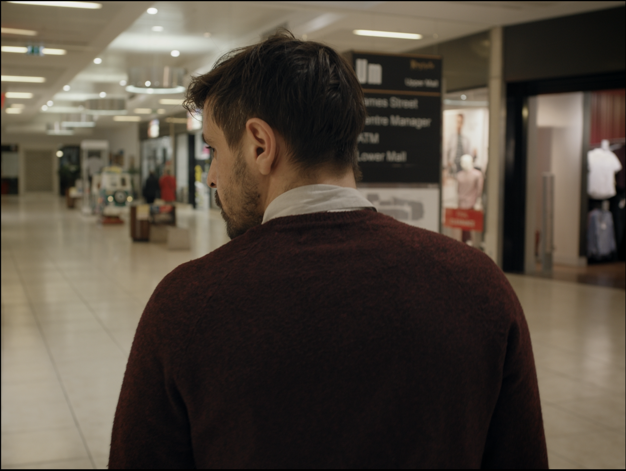 Side profile of a man with dark hair and a beard, wearing a red sweater, in a shopping mall.