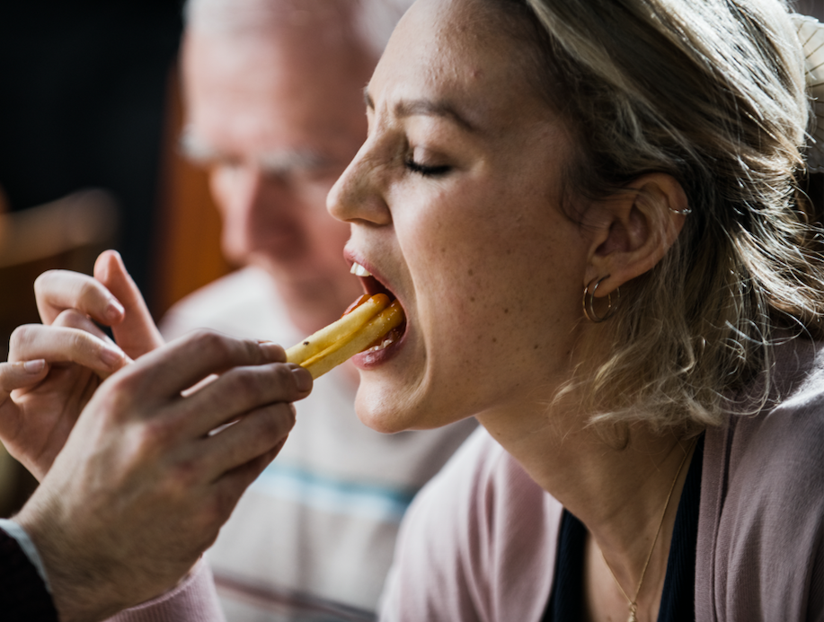 A woman with blonde hair and piercings being fed a French fry by another person in a close-up shot.