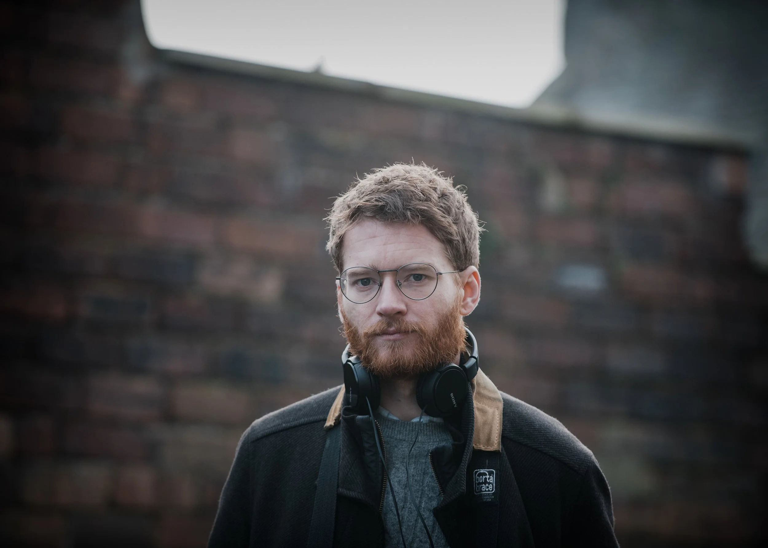 A young man with glasses, red hair, and a beard, wearing headphones around his neck and a dark jacket, standing outdoors against a blurred background of a building or wall.