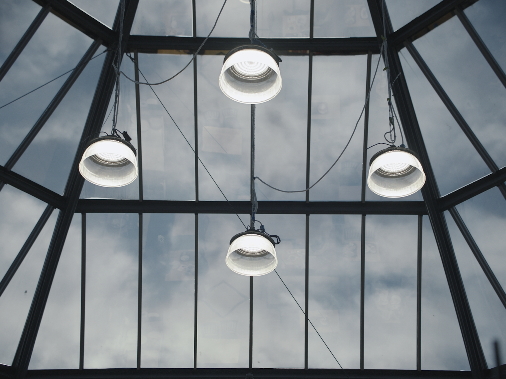Looking up through a glass ceiling with four hanging bright white lights and dark metal framing, against a cloudy sky.