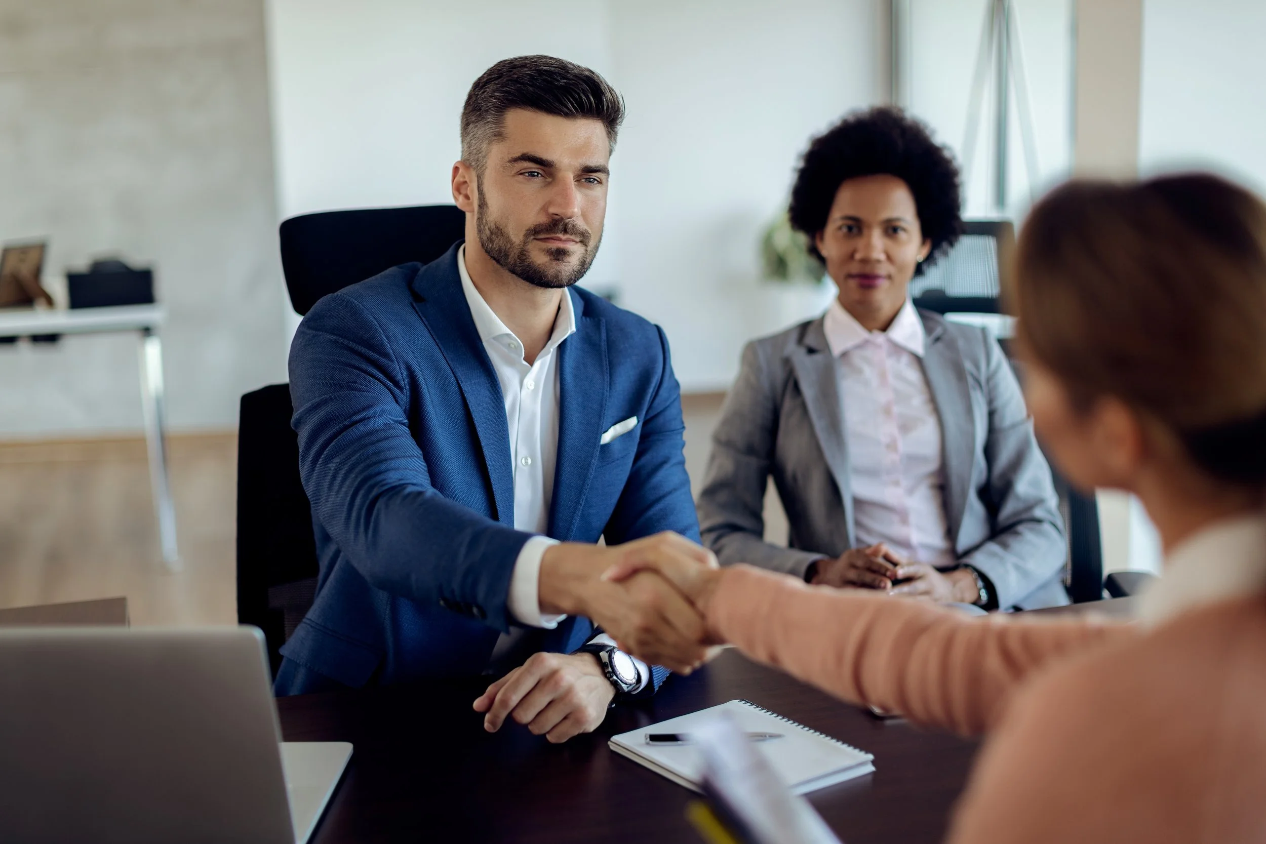 A group of four diverse professionals in a business meeting. The focus is on a man in a blue suit shaking hands with someone off-camera. Behind him, a woman with curly black hair and a grey blazer observes. The setting appears to be a modern office with natural lighting.