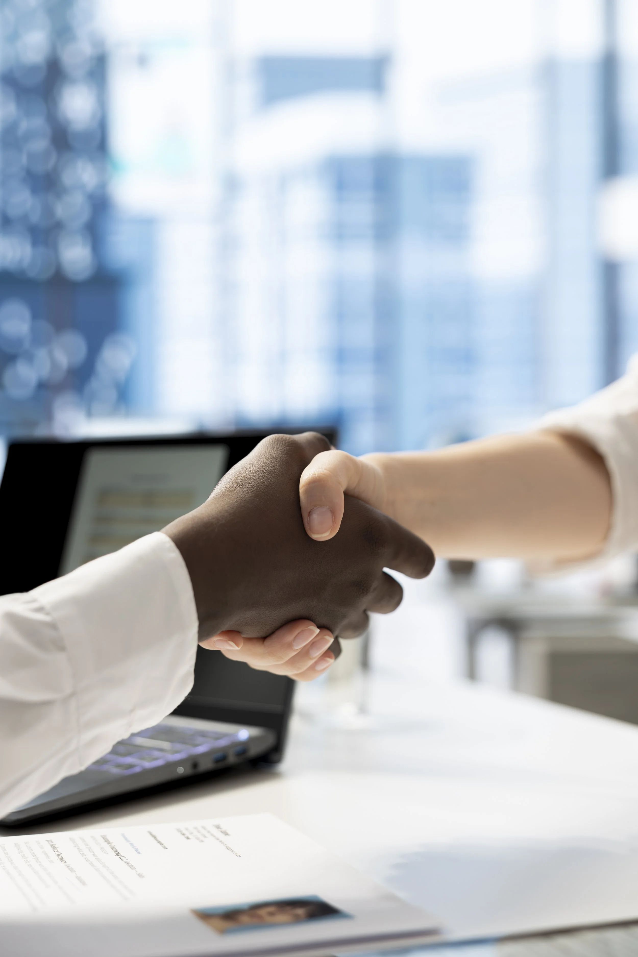 Two people shaking hands over a desk, with a laptop and documents on the table, in an office setting.