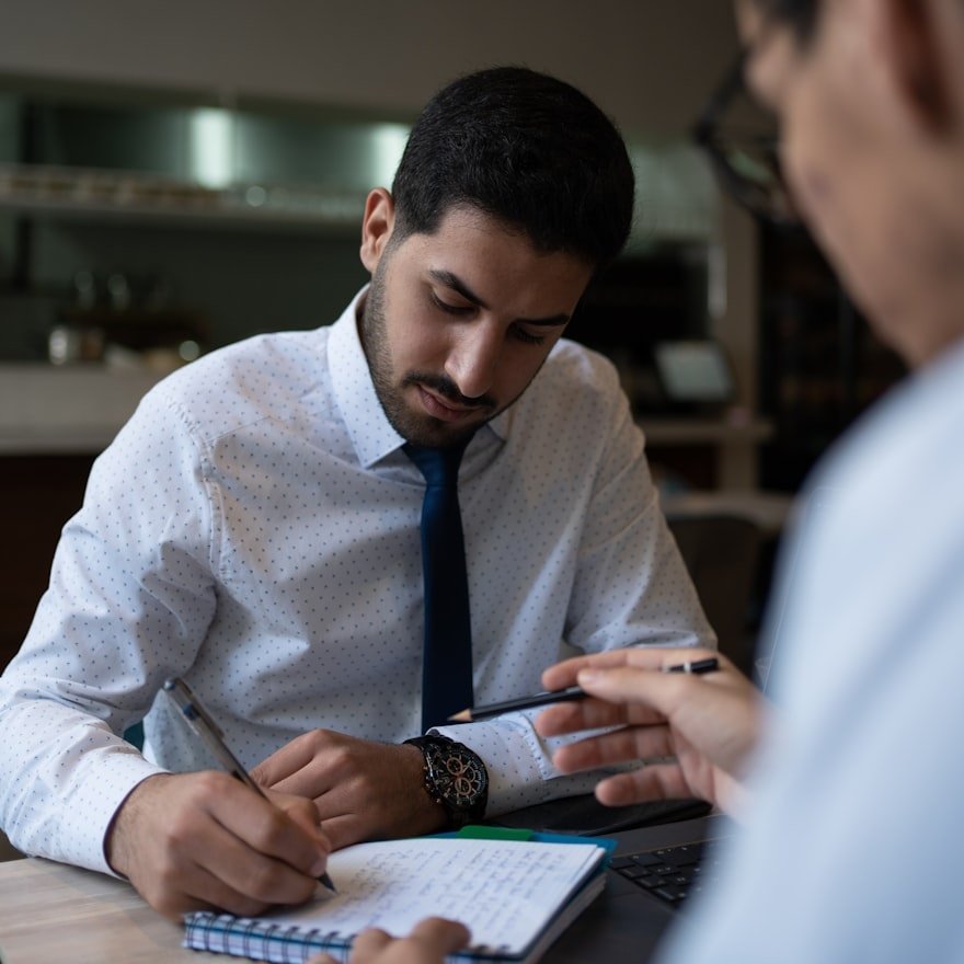 Two men engaged in a discussion, one taking notes and the other holding a phone, in an office or conference room setting.