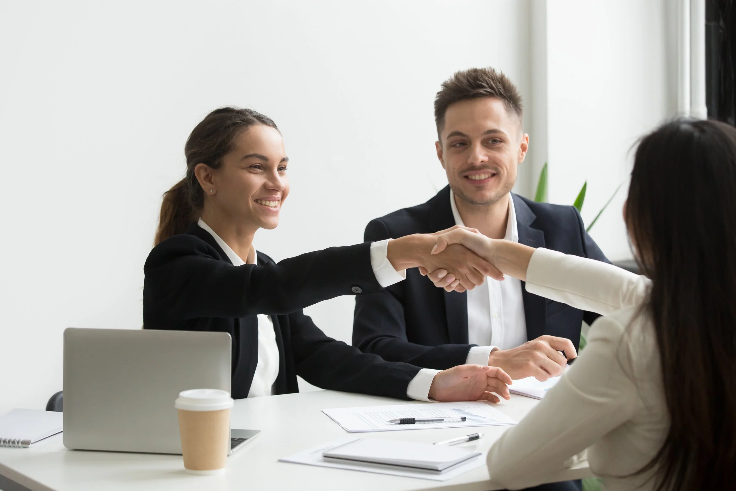 Business meeting with diverse professionals, two women and a man, exchanging a handshake in a modern office setting with a coffee cup and documents on the table.