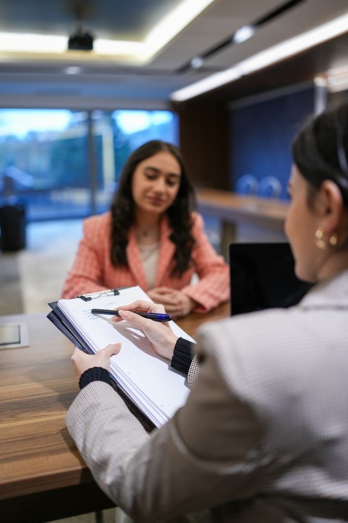 Two women having a job interview in a modern office setting. One woman is sitting across the table, and the other is taking notes on a notepad.