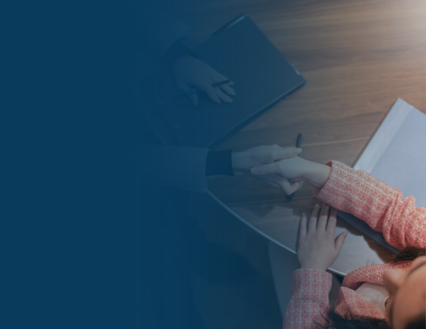 Two people shaking hands over a wooden table during a business meeting, with a notebook and a pen on the table.