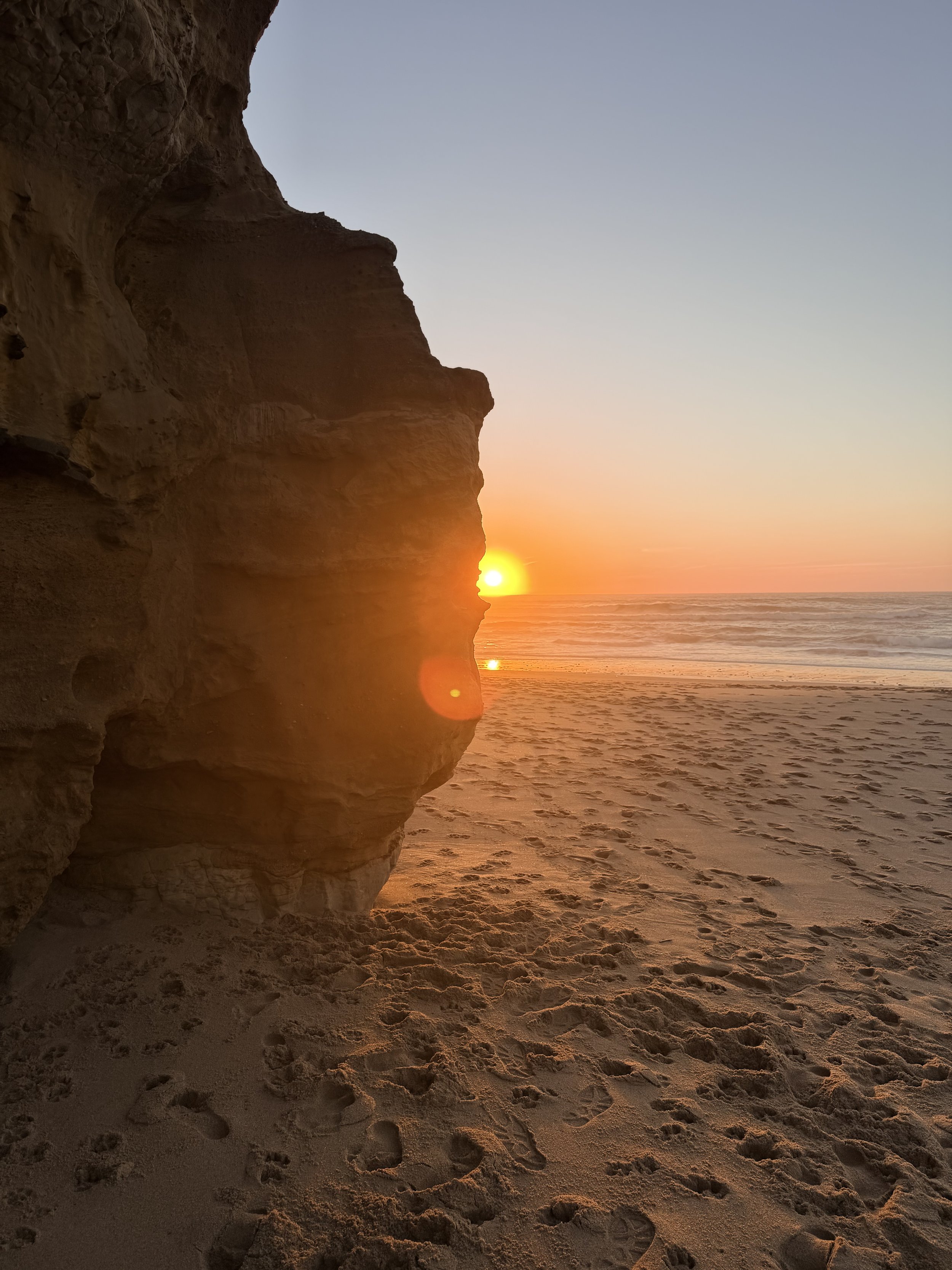 View of a beach at sunset with sand, ocean waves, and a large rock formation on the left side.