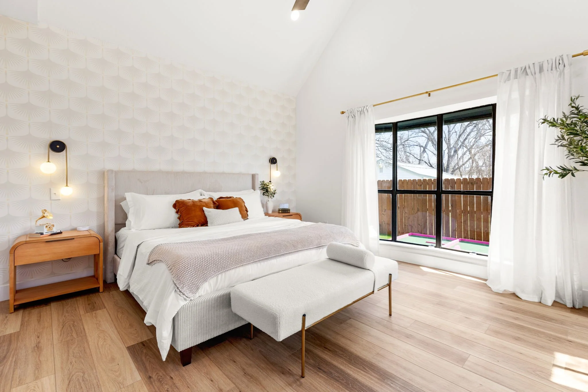 A bedroom with a large bed with white linens and rust-colored pillows, two black wall-mounted lamps on either side, a wooden nightstand with a small plant, and a window with white curtains showing a backyard with trees and a fence.