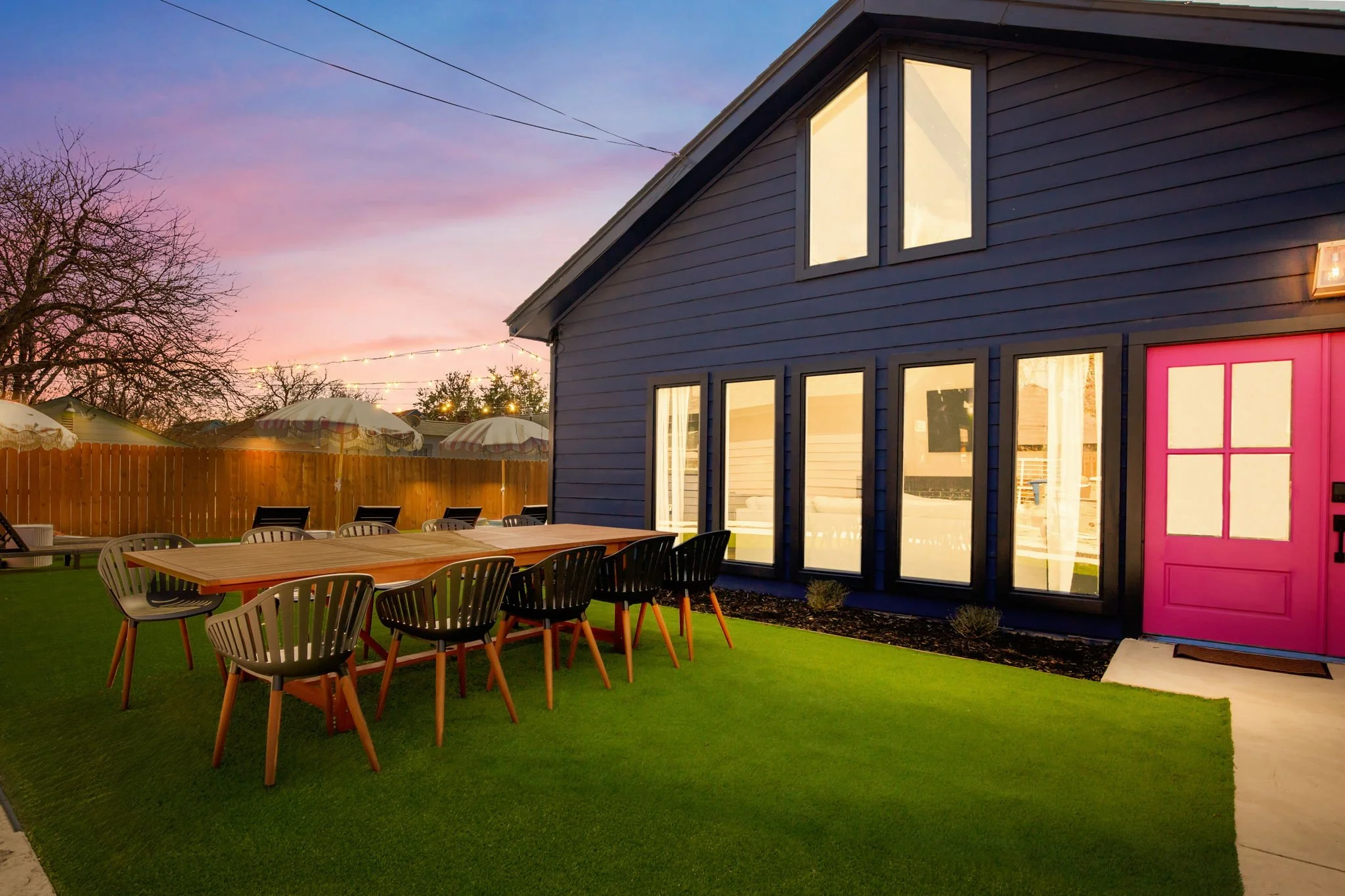 A backyard patio scene at sunset featuring a blue house with large windows and a pink door. There is a wooden table surrounded by black chairs on a green artificial grass surface, with a fence, trees, and umbrellas in the background and string lights
