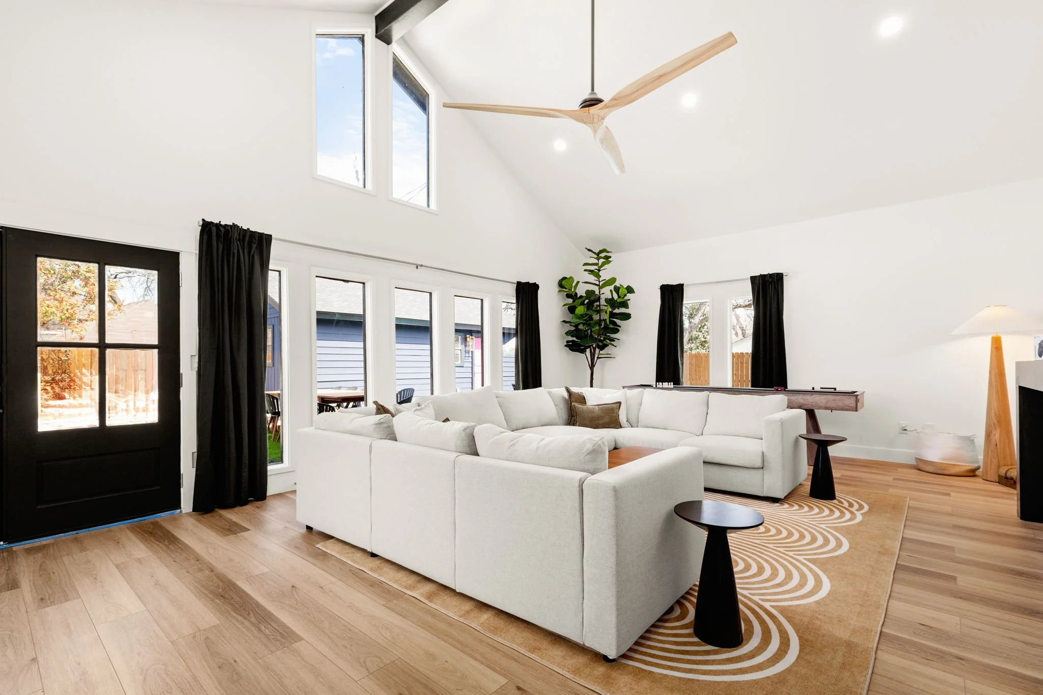 Living room with white L-shaped sofa, black side tables, wooden flooring, large windows with black curtains, a ceiling fan, a standing lamp, and a tall green houseplant.