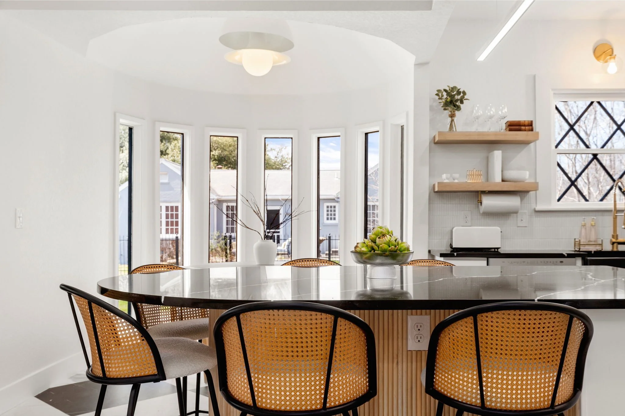 A modern kitchen and dining area with a round white table, beige chairs, a bowl of artichokes, and large windows showing outdoor trees and houses; minimalistic decor with floating shelves, plants, and wine glasses.