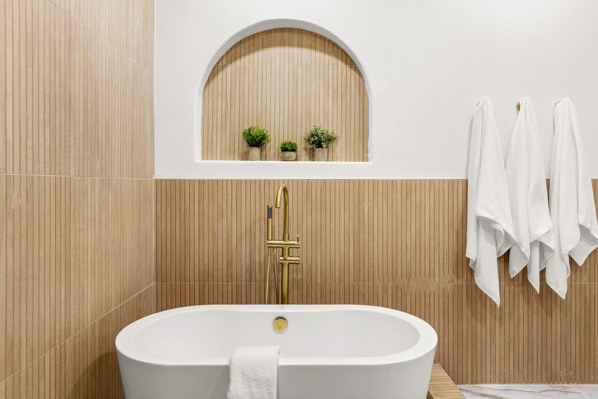 A modern bathtub with gold fixtures surrounded by wood-paneled walls and white towels hanging on hooks next to a white wall with a semi-circular wood accent and potted plants.