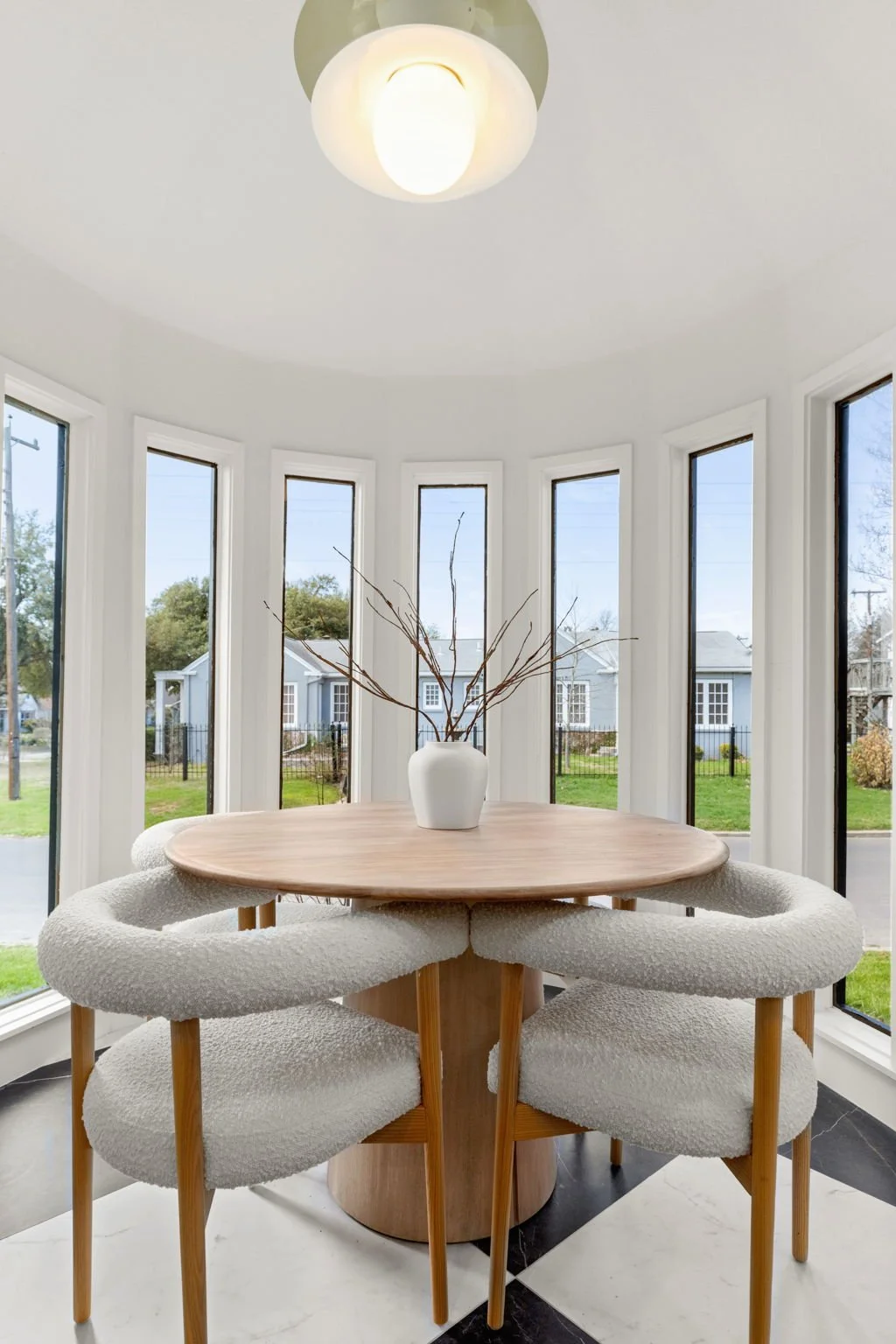 Bright dining nook with a round wooden table and four white textured chairs, positioned in front of large windows overlooking a suburban street, with a ceiling light fixture above and a white vase with branches as a centerpiece.