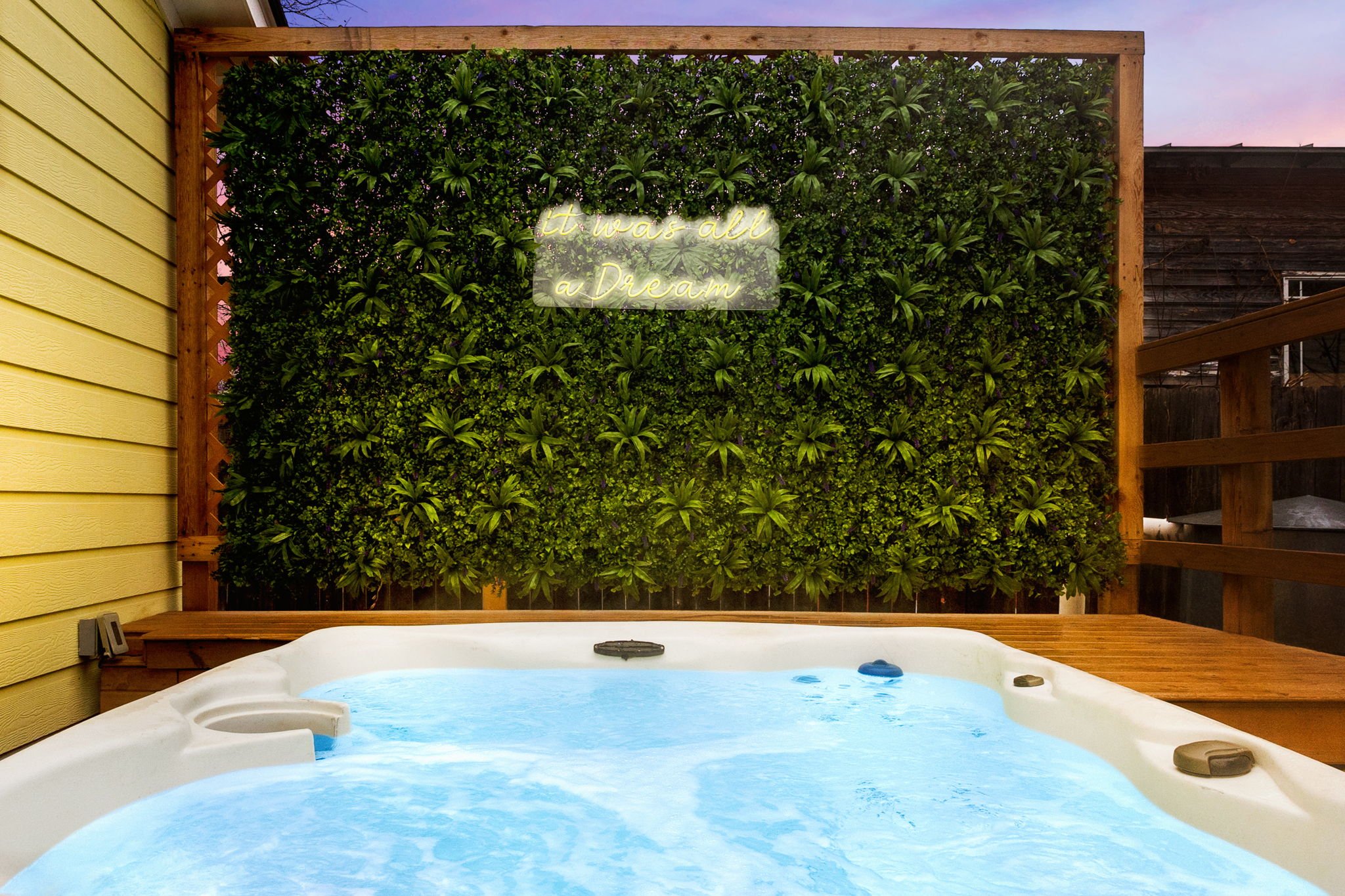 A backyard hot tub with blue water, situated in front of a lush green vertical garden with a neon sign that reads 'it was all a Dream,' attached to a wooden frame. To the left, there is yellow siding on the house, and above, the sky appears to be at dusk.