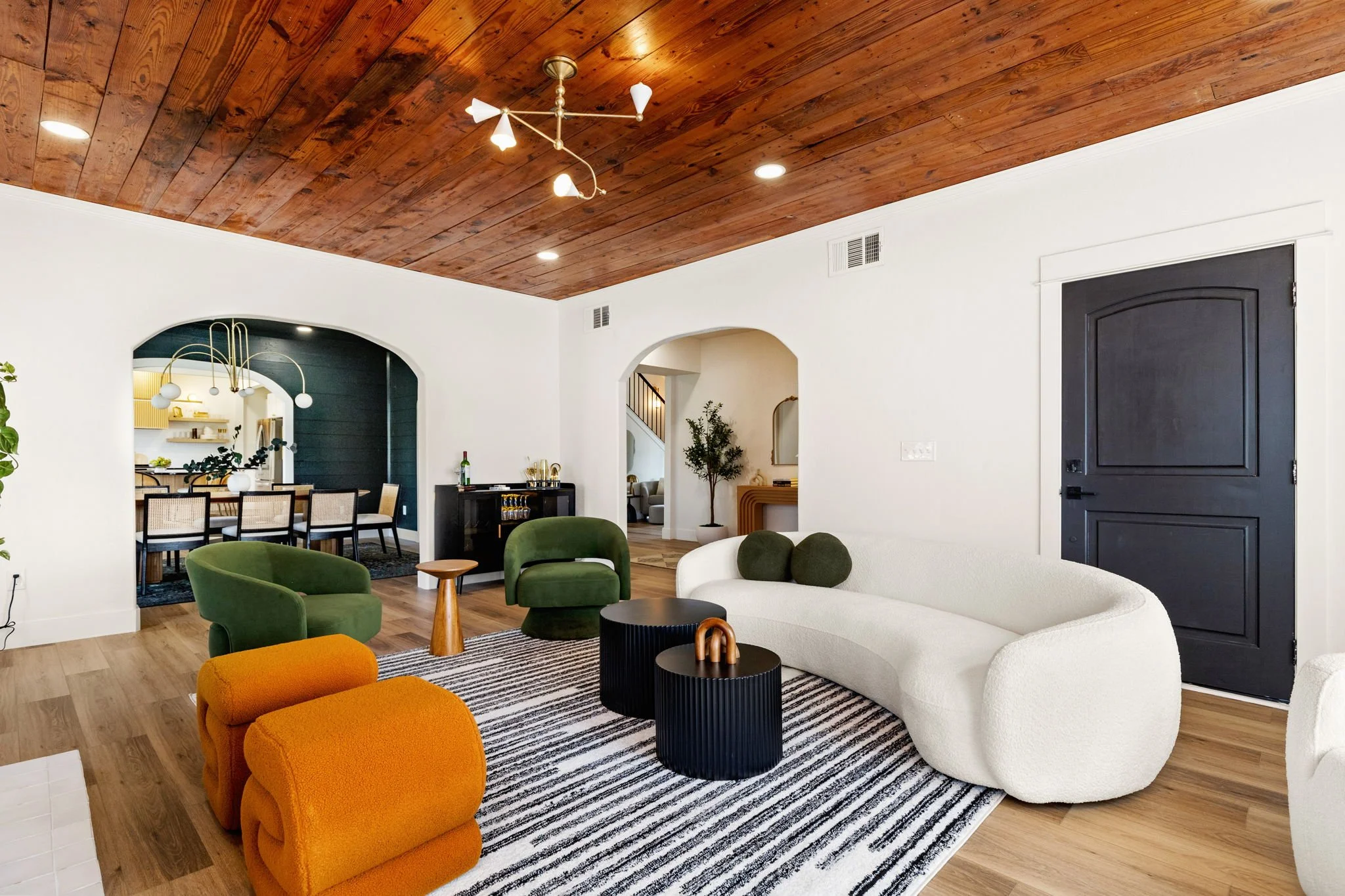 Bright living room with white walls, a wooden ceiling, and a mix of modern furniture including green and orange chairs, a white sofa, and black side tables. A striped rug is on the wooden floor, and decorative plants and lights are also visible.