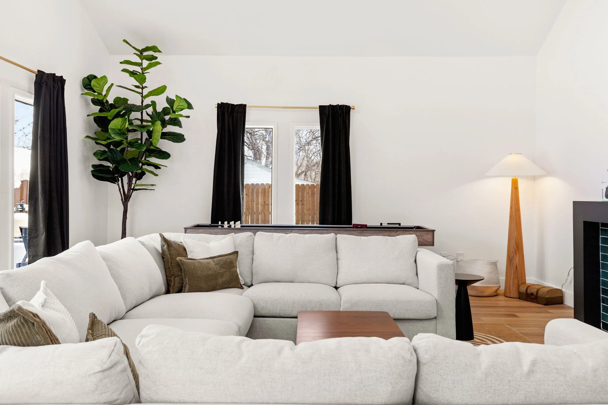 Living room with a large white sectional sofa, brown throw pillows, a tall potted plant, black curtains on windows, a wooden floor lamp on the right, and a small table.