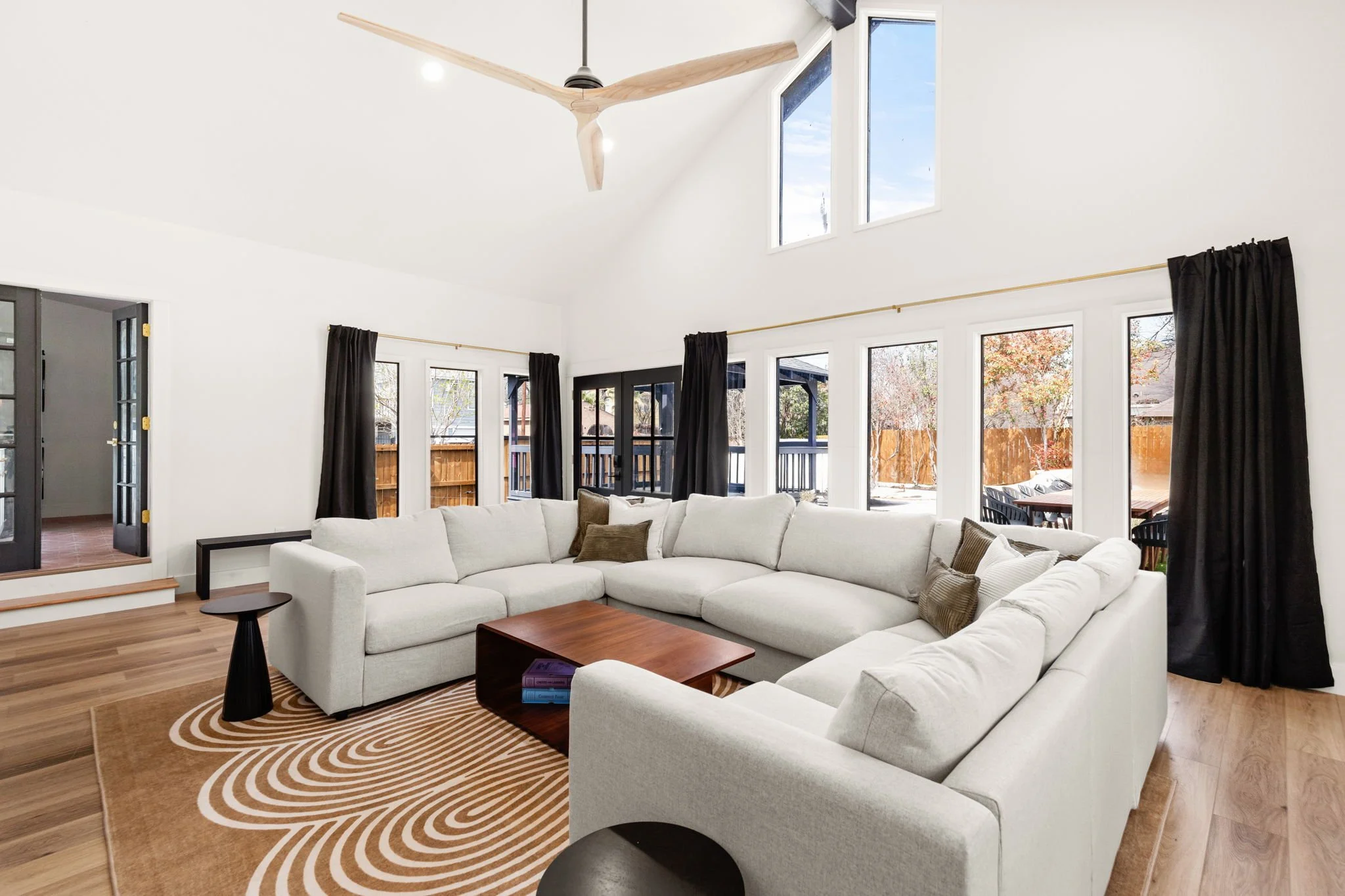 Living room with white sectional sofa, black curtains, wood coffee table, ceiling fan with wooden blades, large windows showing outdoor deck and yard, and a patterned rug.