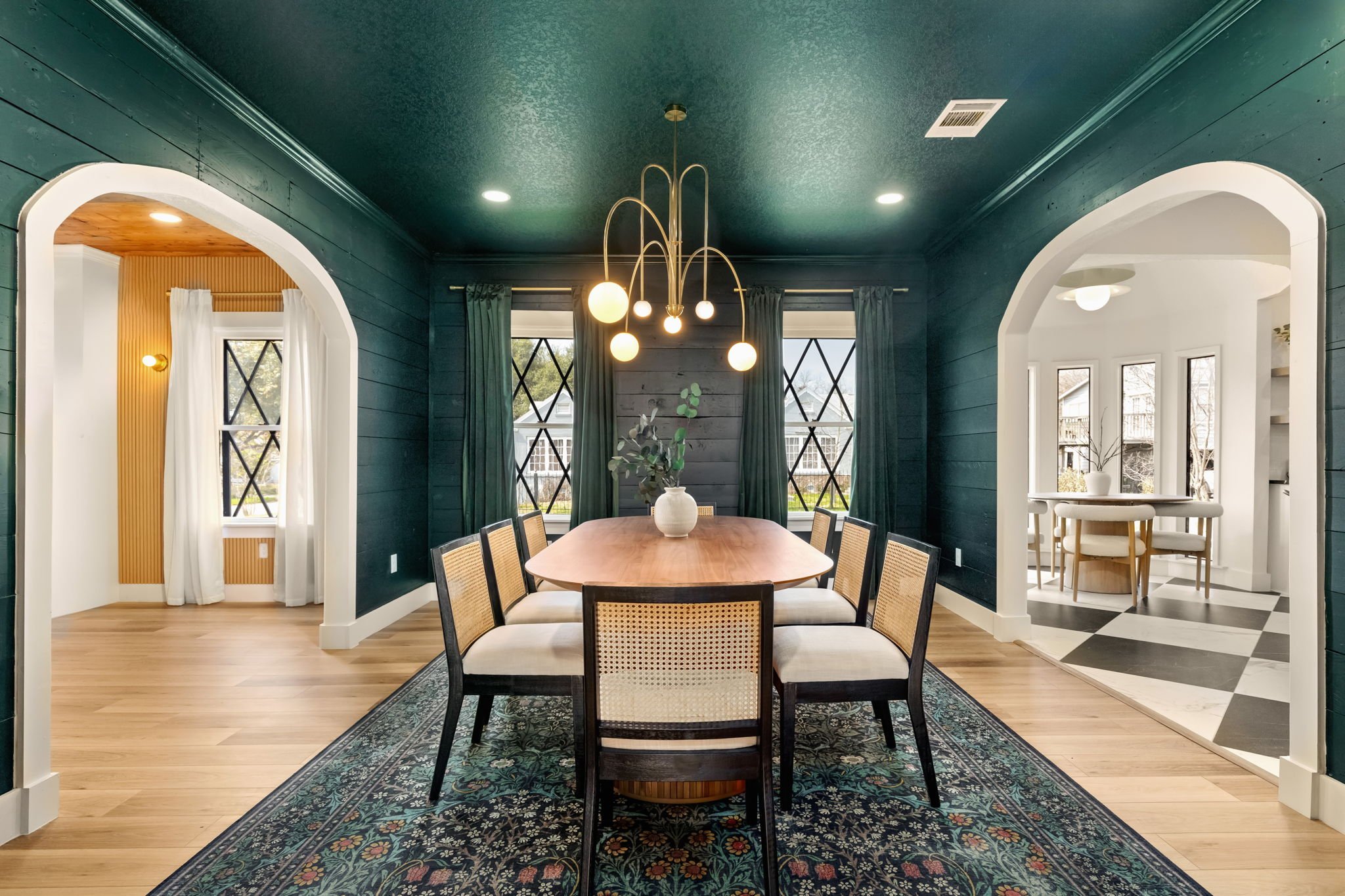 Dining room with blue walls, large windows with green curtains, and a hanging modern chandelier above a wooden dining table surrounded by chairs. There's a patterned rug underneath the table and a vase with greenery in the center.