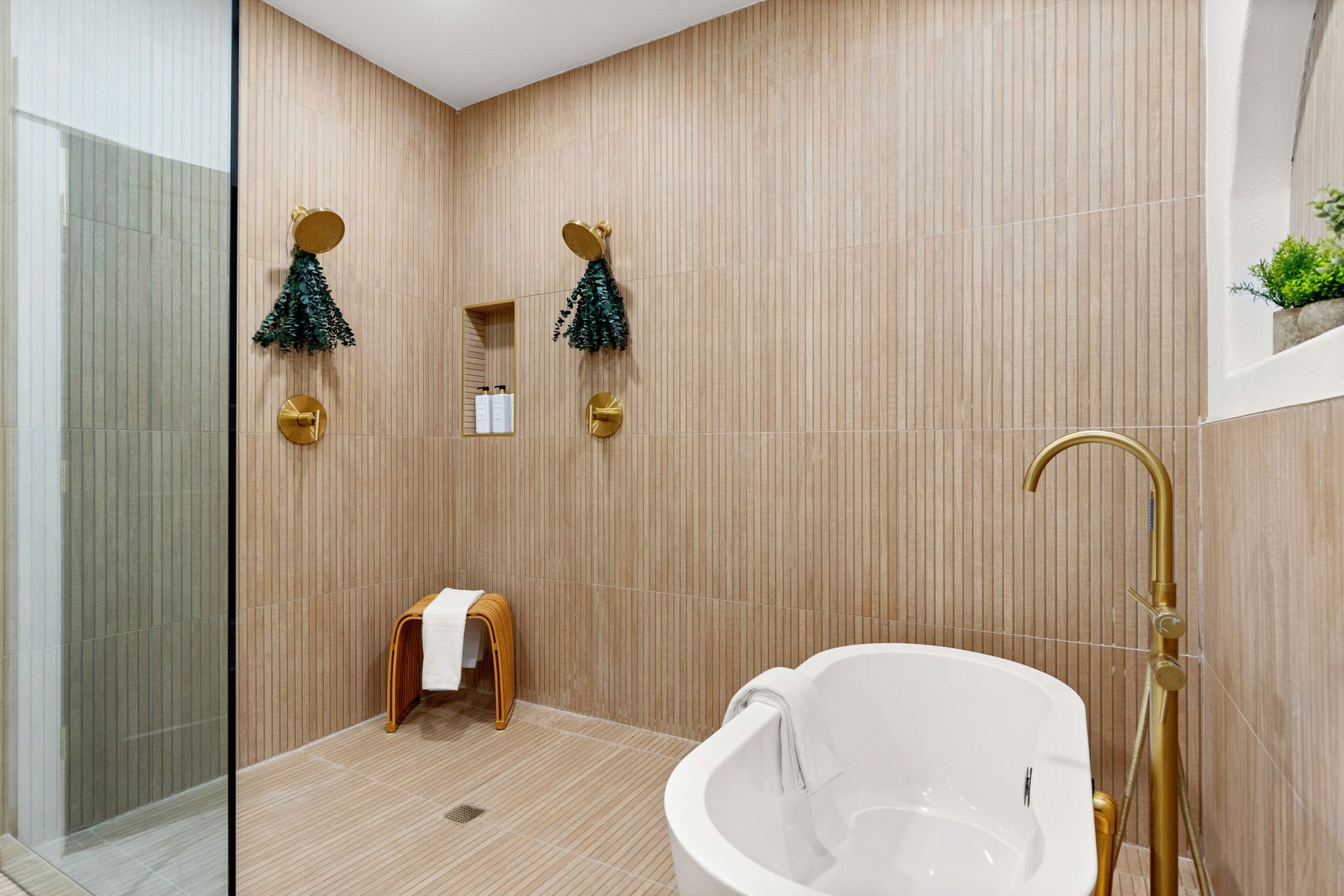 A modern bathroom with wood-textured tiles on walls and floor, featuring a walk-in shower with a glass door, a white bathtub with a rolled towel, gold fixtures, and decorative greenery hanging on the wall.