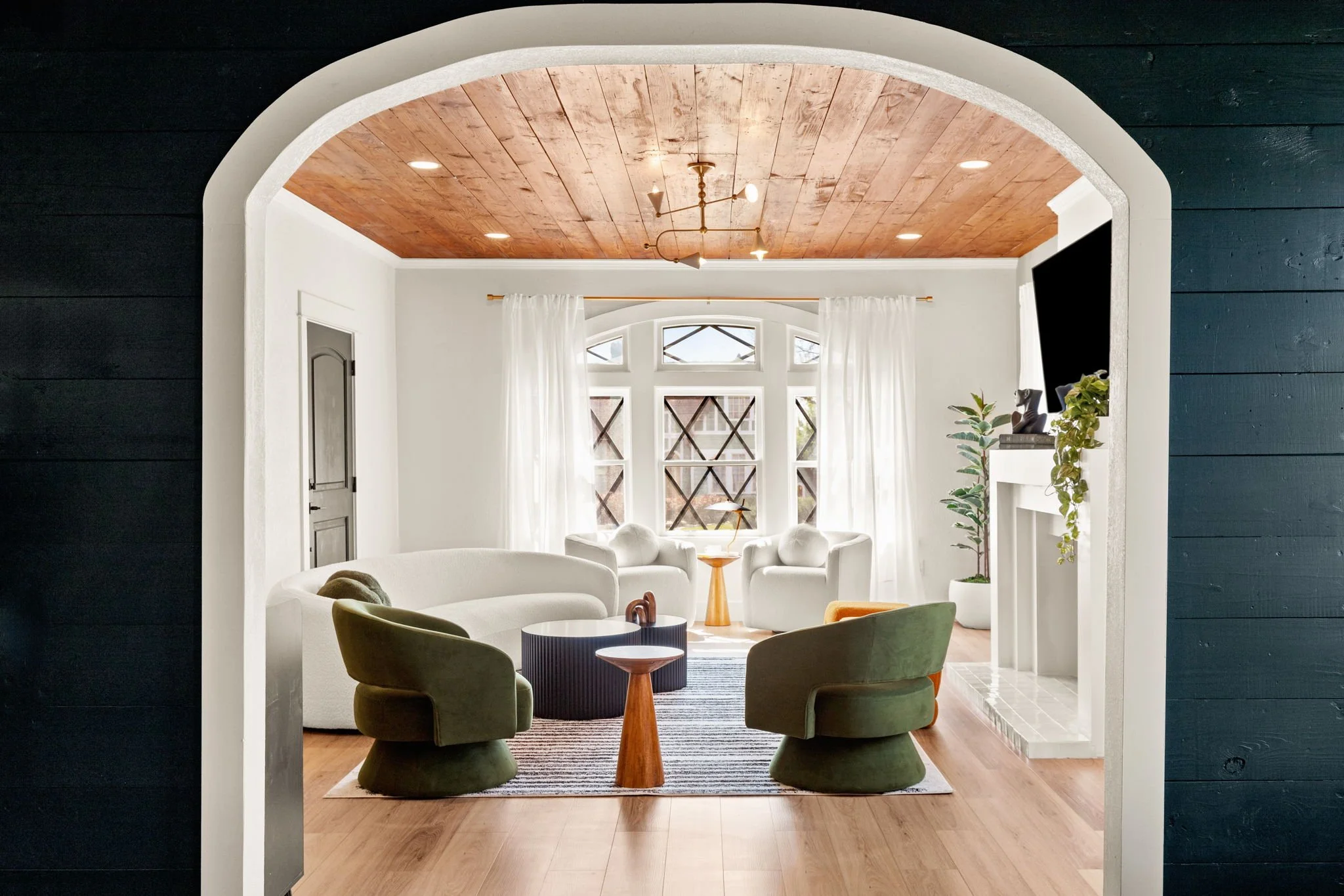 Living room with white walls and large windows, wooden ceiling, and modern furniture including green armchairs, a white sofa, and a black coffee table, decorated with plants and a wall-mounted TV.