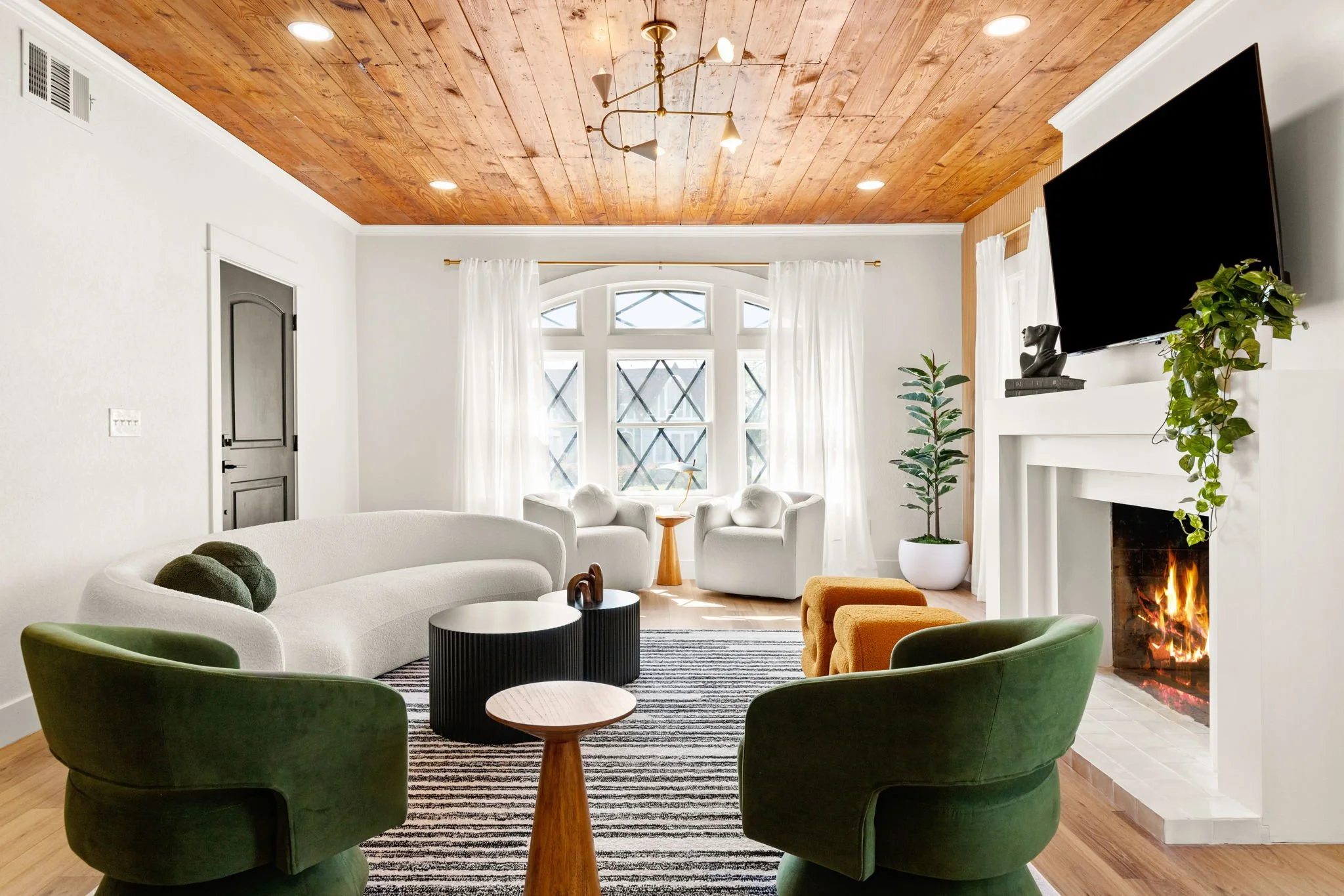 A living room with a wooden ceiling, white walls, and large window with sheer curtains. There are white chairs, a green curved sofa, and a fireplace with a TV mounted above it. Decor includes a plant and a sculpture.
