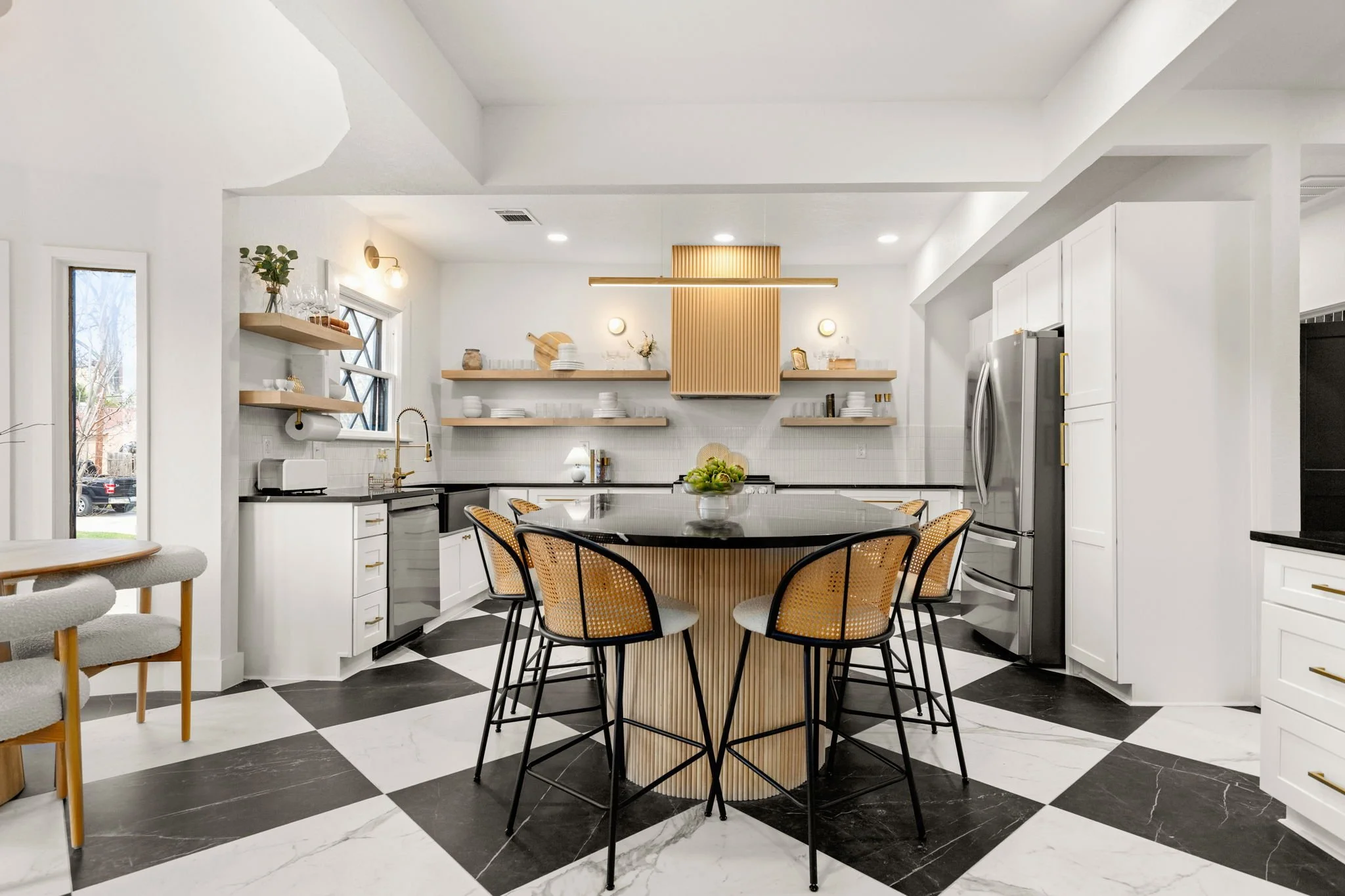 Modern kitchen and dining area with black and white checkered marble floor, white cabinets, open wooden shelves, black round table, and woven back chairs. Light fixtures and decorative items are on the shelves, with windows letting in natural light.