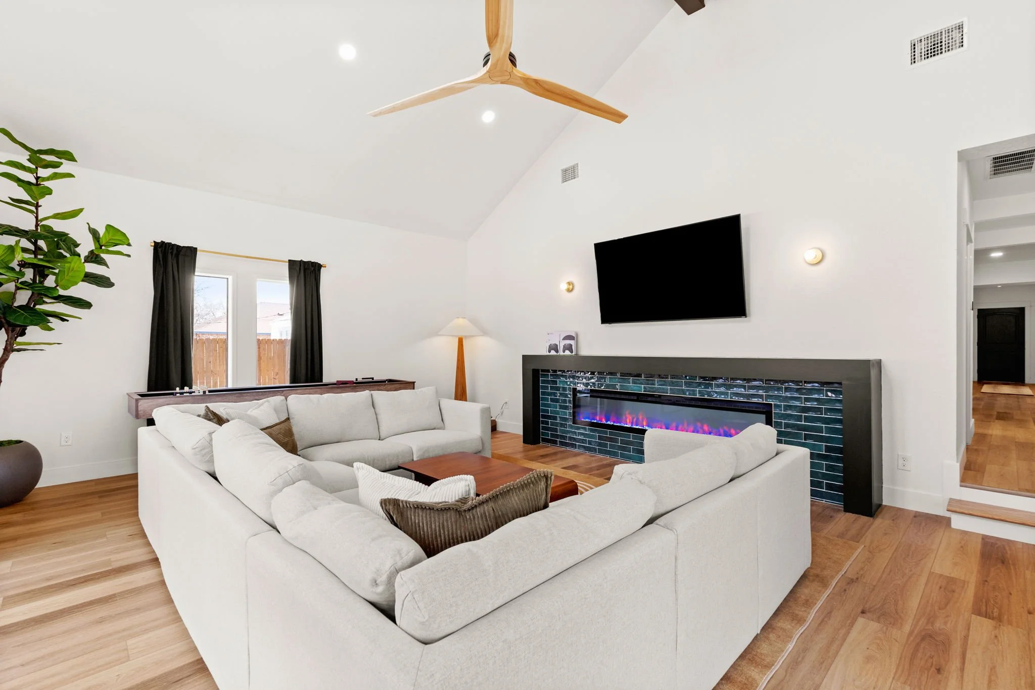 Spacious living room with white sectional sofa, wooden coffee table, black fireplace with colorful flames, large flat-screen TV, potted plant, and ceiling fan, with windows and black curtains
