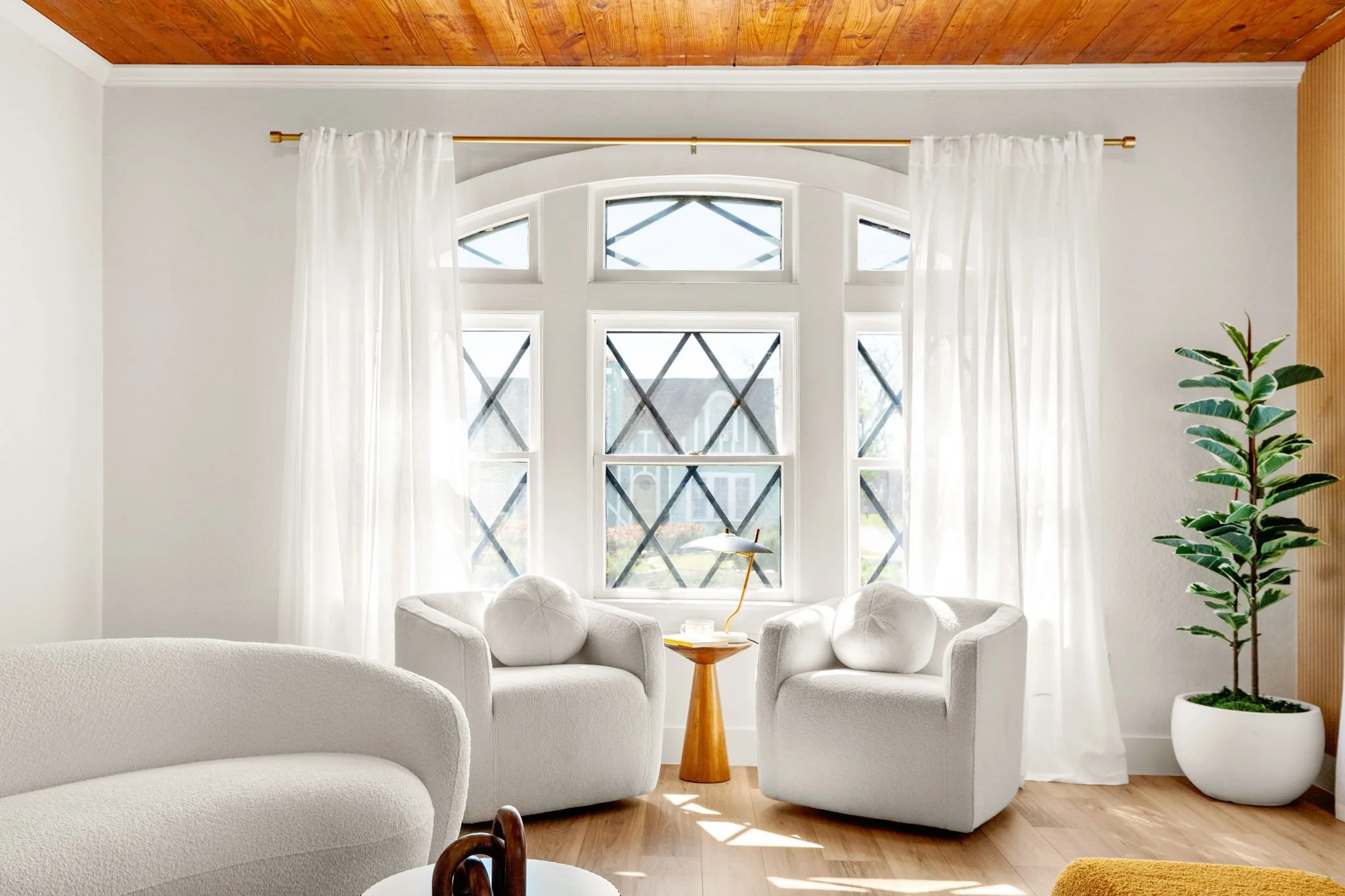 Living room with white upholstered furniture arranged around a wooden side table, large arched window with multiple panes, sheer white curtains, wooden ceiling, and a tall potted plant on the right.