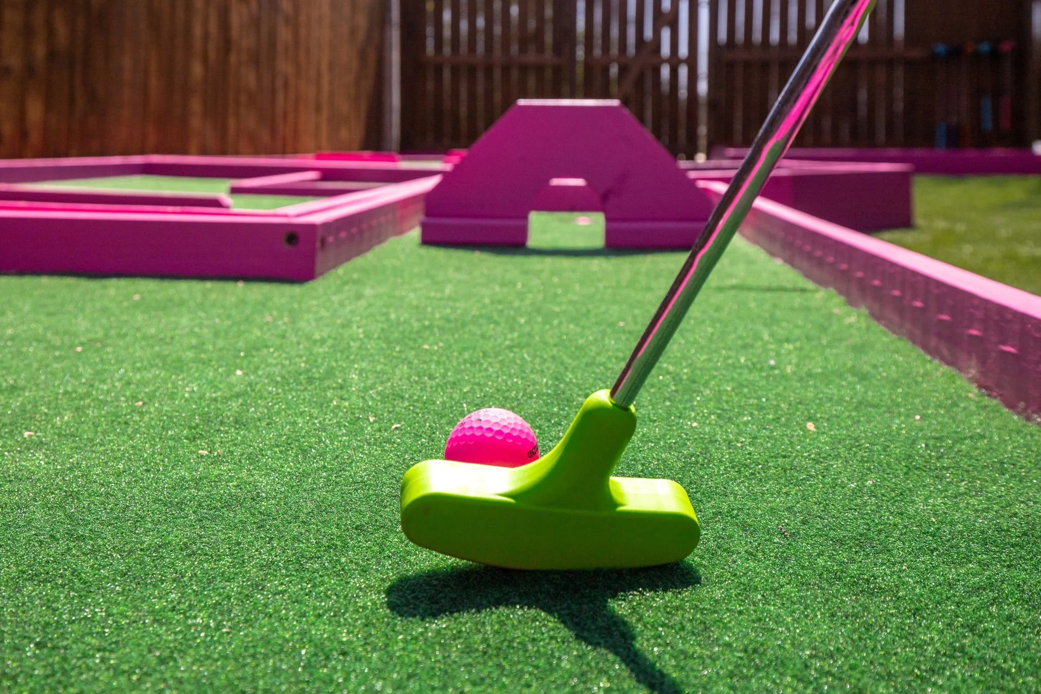 A miniature golf course with a pink obstacle and green artificial turf, a pink golf ball, and a yellow plastic golf tee, with a wooden fence background.