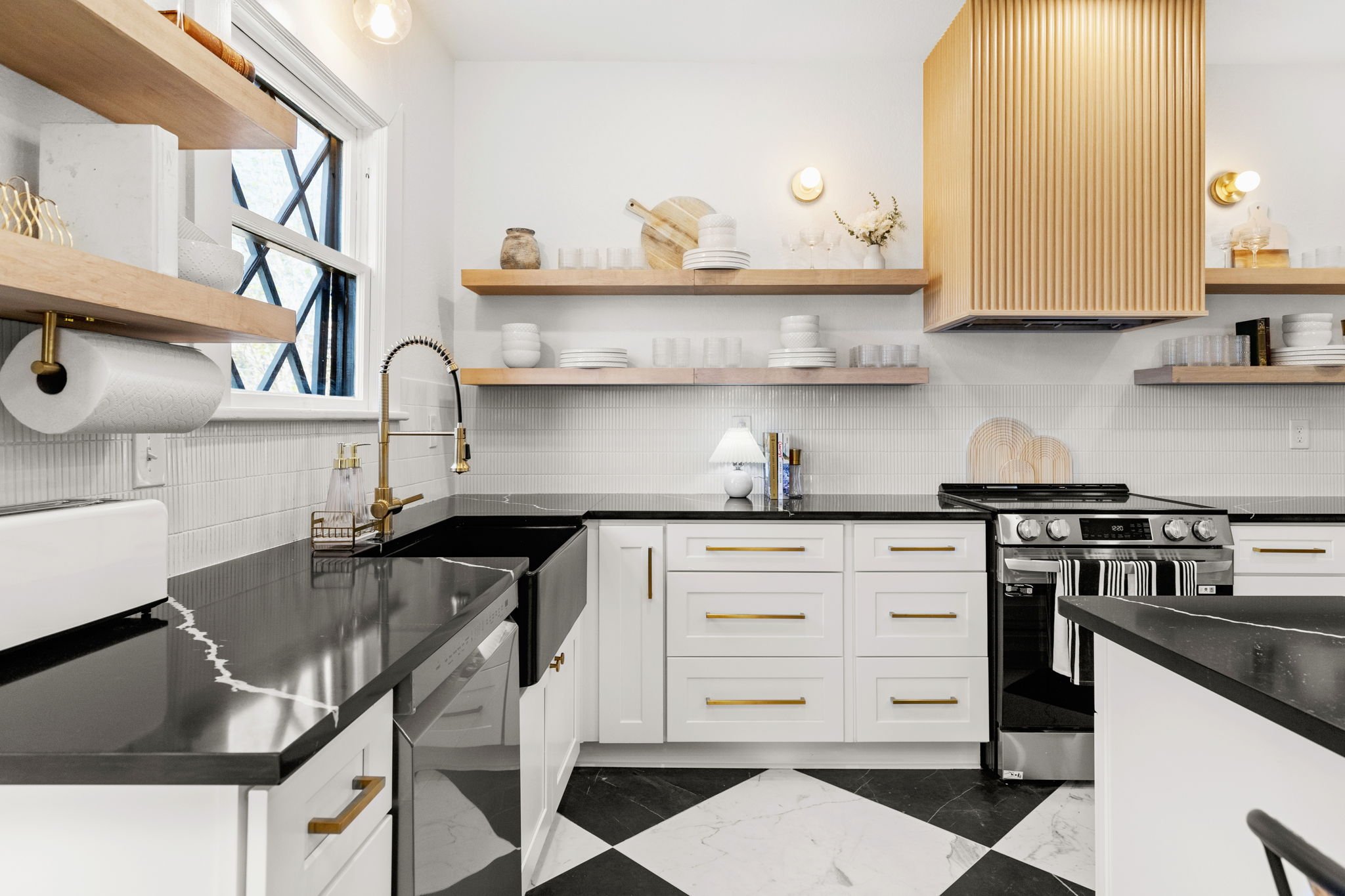 Modern kitchen with white cabinets, black countertops, and gold hardware. Open wooden shelves hold white dishes and vases. There is a large window, a stove, and a small lamp on the counter.