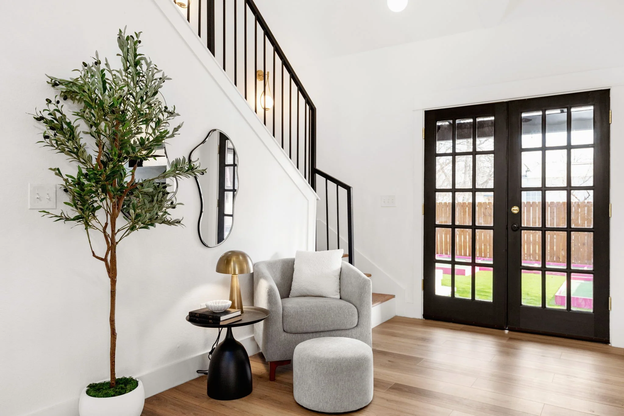 Living room corner with gray armchair, matching ottoman, small black side table with books and decorative objects, tall potted plant, black-framed glass double doors leading to backyard, white walls, wood flooring, and a staircase with black railing.
