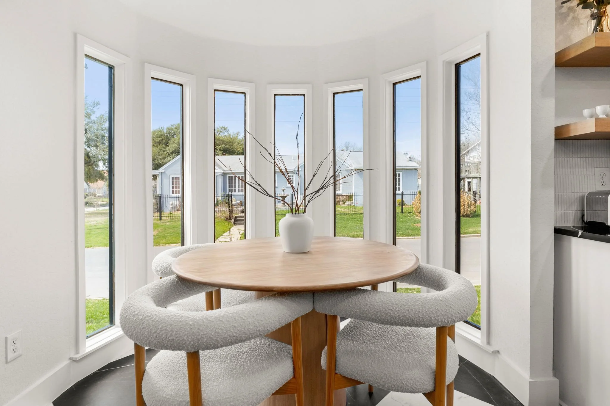 A dining area with a round wooden table and four upholstered chairs, a white vase with decorative branches, large windows with a backyard view, and a kitchen corner on the right.