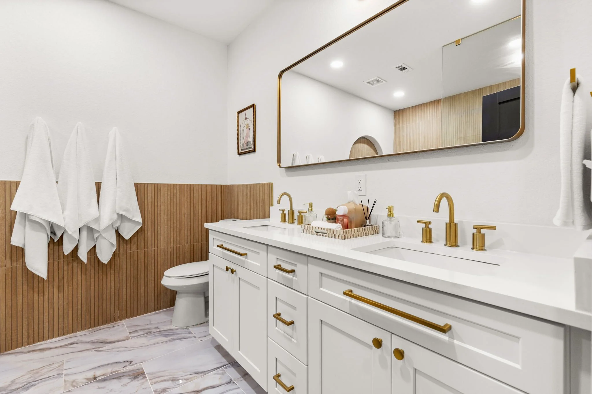 Modern bathroom with white vanity, gold fixtures, large mirror, and wood-paneled wall with towels hanging on hooks.