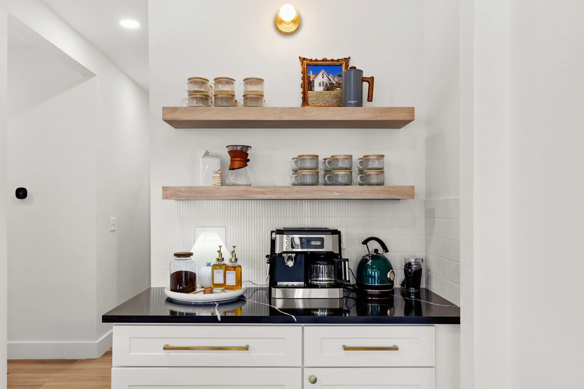 Kitchen countertop with coffee machine, kettle, and jars, two wooden shelves with coffee mugs and a framed picture of a house on the wall.