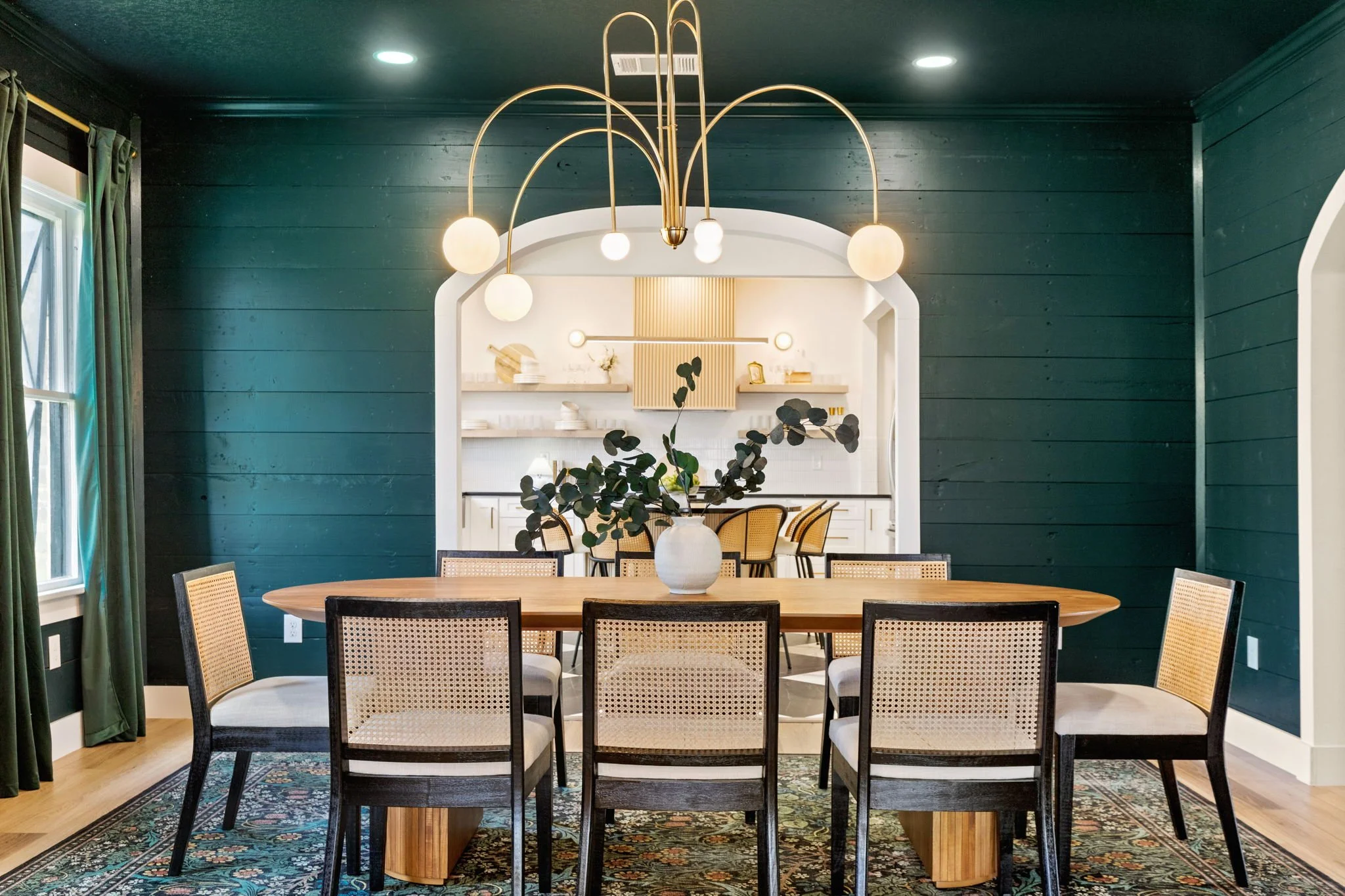 Interior of a dining room with a wooden table, eight chairs with woven seats, a rug, a large vase with eucalyptus on the table, green curtains, a pendant light fixture with multiple bulbs, dark teal paneled walls, and a view into a bright kitchen are