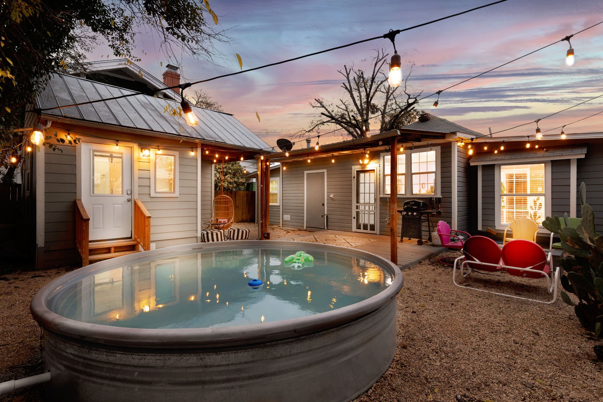 A backyard sunset scene with a above ground pool, string lights hanging above, and a patio with outdoor furniture. There are houses with windows and trees in the background.