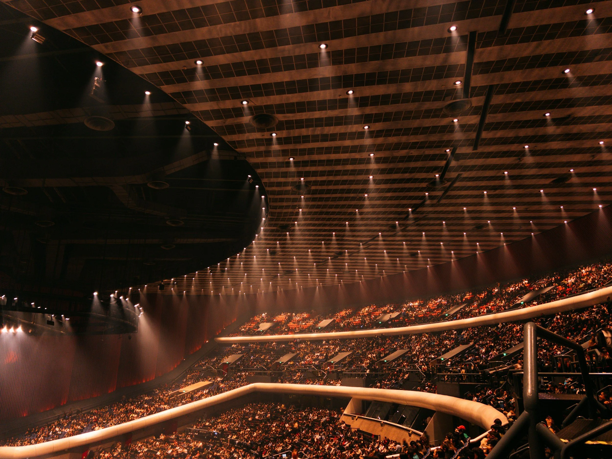 Interior of a large, modern auditorium filled with people, featuring a high curved ceiling with numerous ceiling lights and a stage at the front.