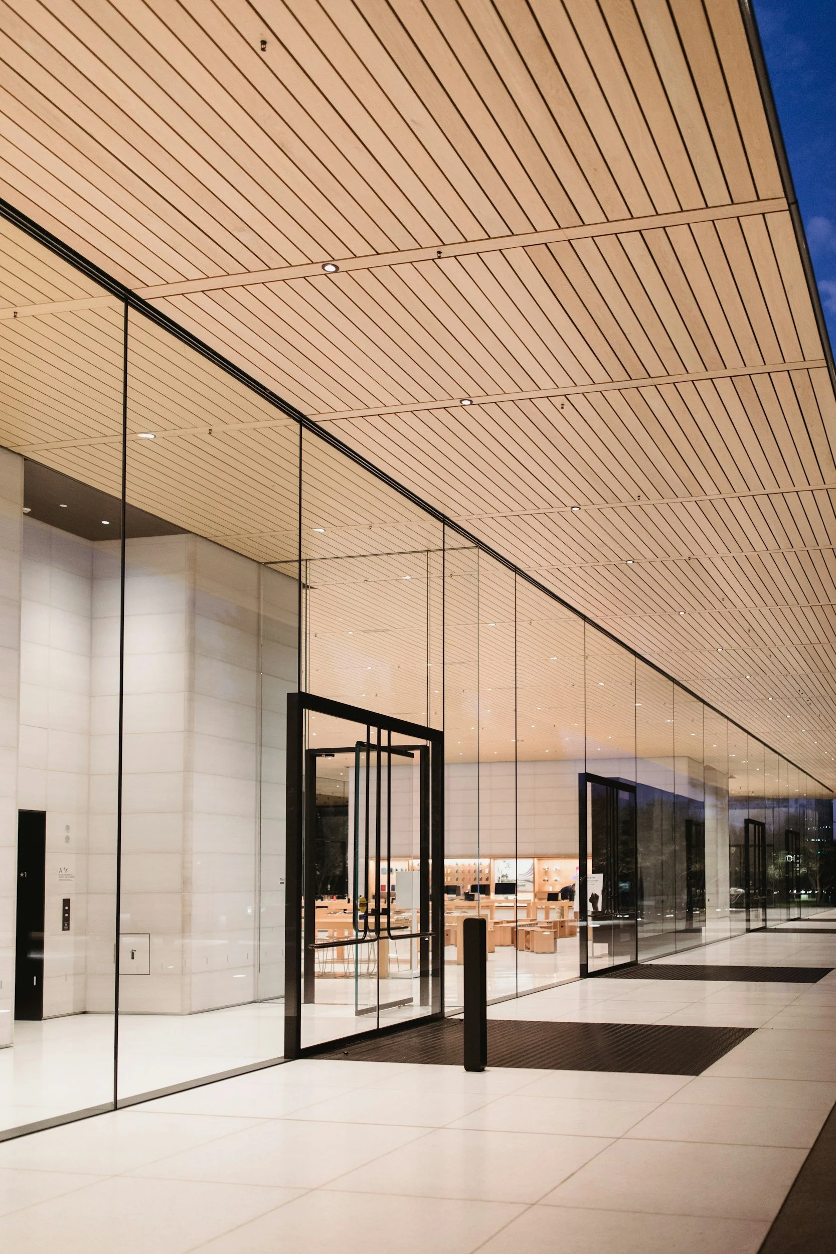 The image shows the front entrance of a modern building with glass walls and a wooden ceiling, with a glimpse of the interior visible through the glass.