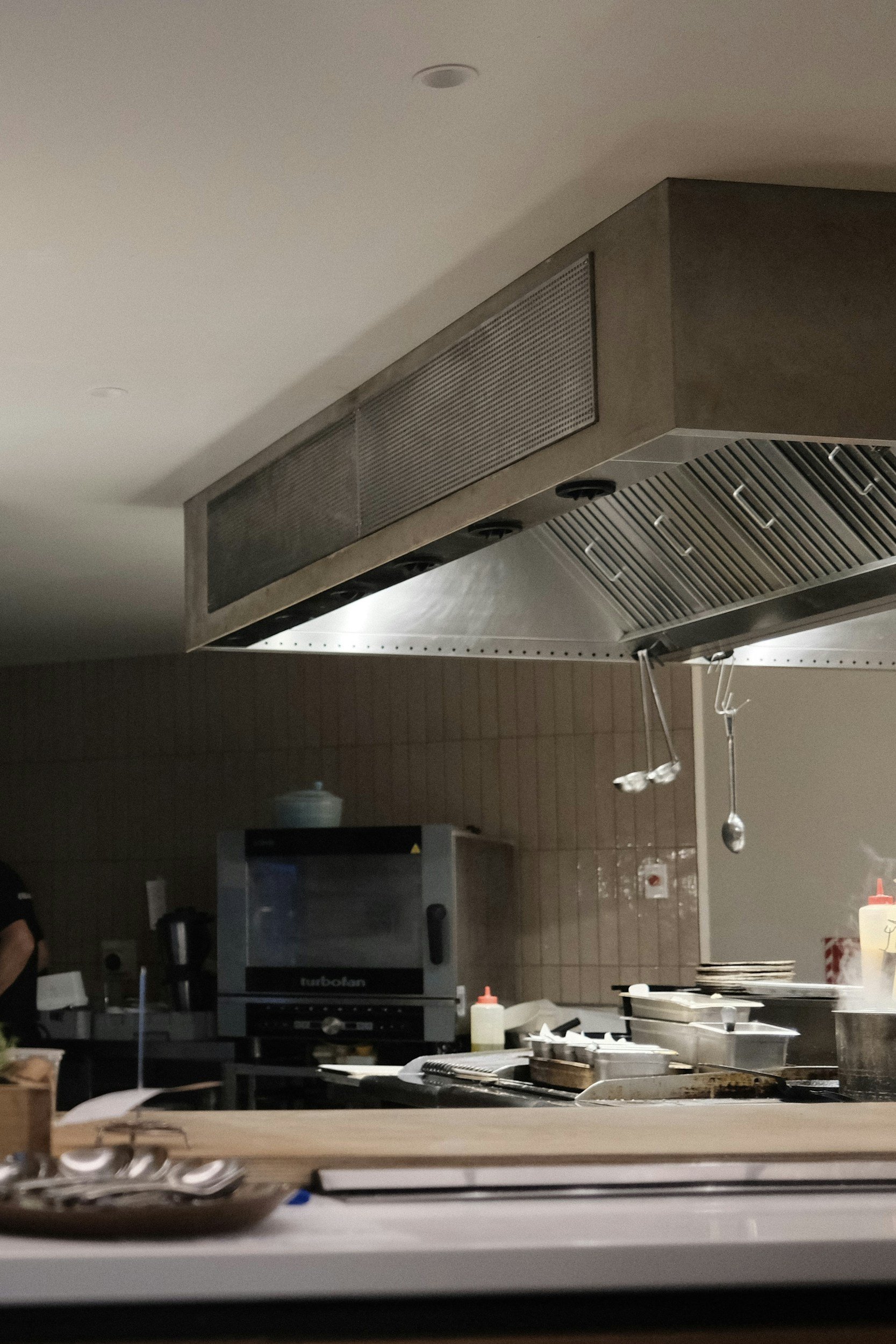 View of a commercial kitchen with a stainless steel range hood, a microwave oven, open shelves with dishes, and cooking utensils on the countertop.