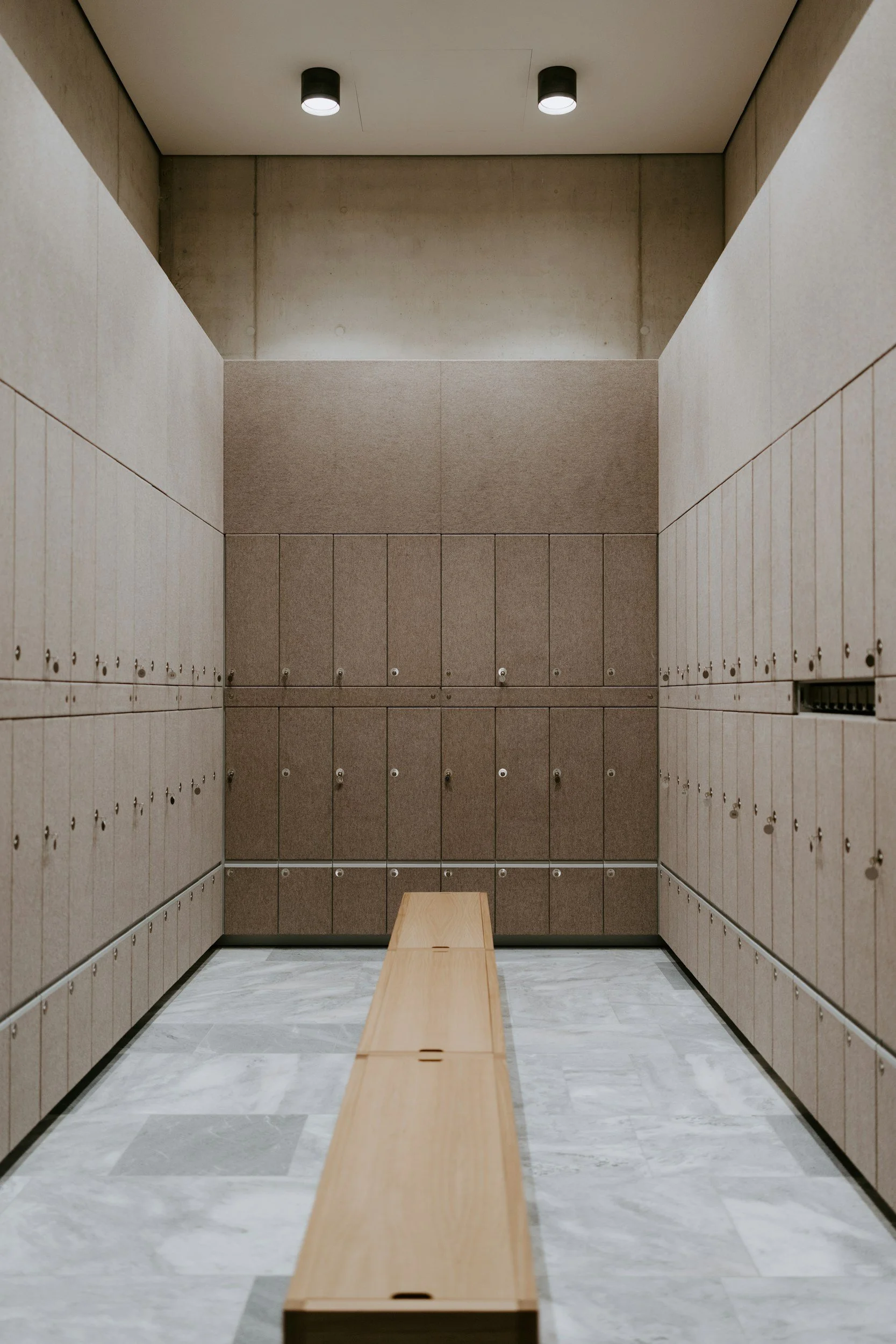 Locker room with beige lockers on both sides, a wooden bench in the center, and ceiling lights.