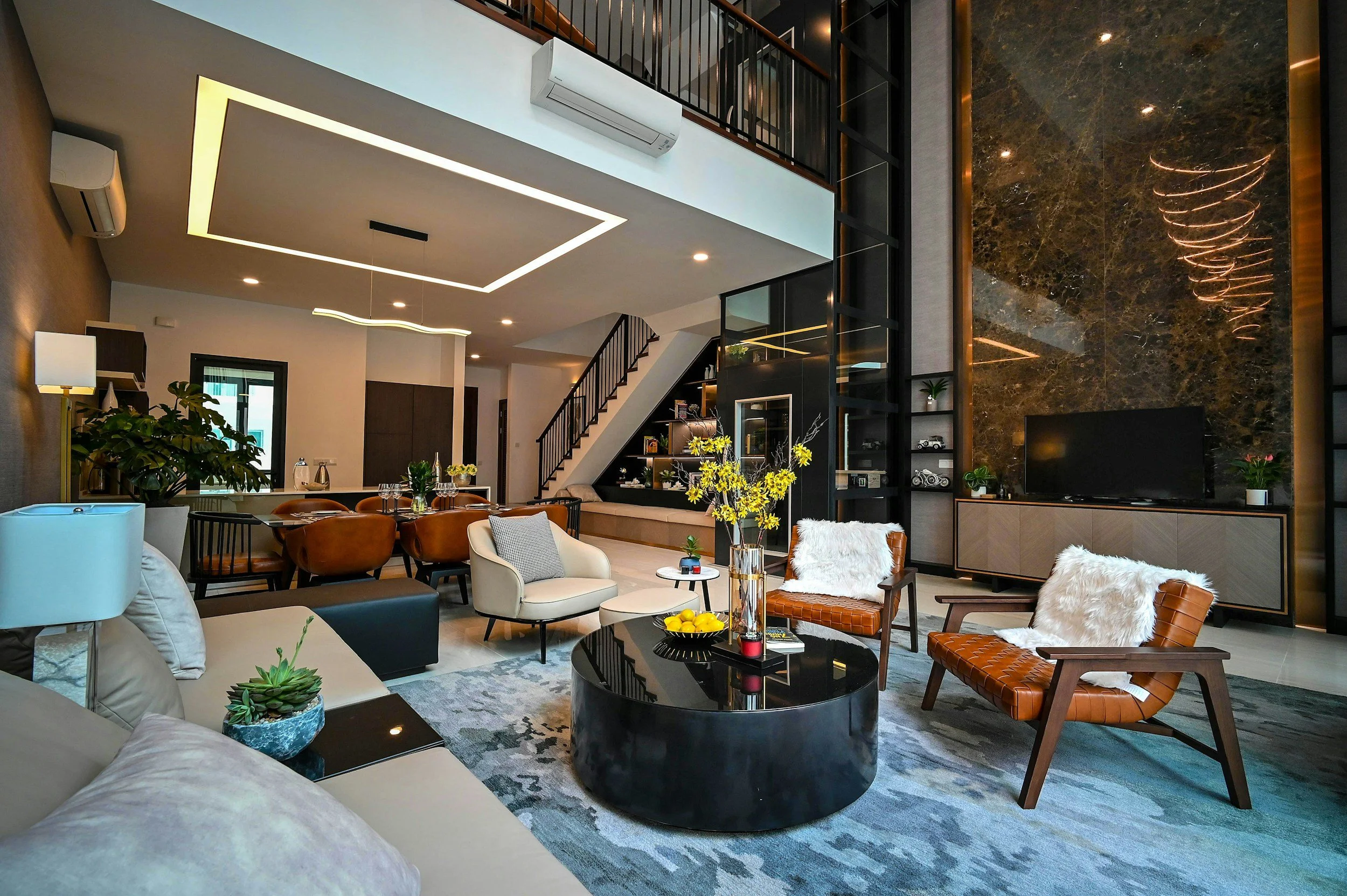 Modern living room with high ceiling featuring ceiling lighting, staircase with black railing, large marbled wall behind a flat-screen TV, and various chairs and sofas decorated with cushions and ties of yellow flowers.